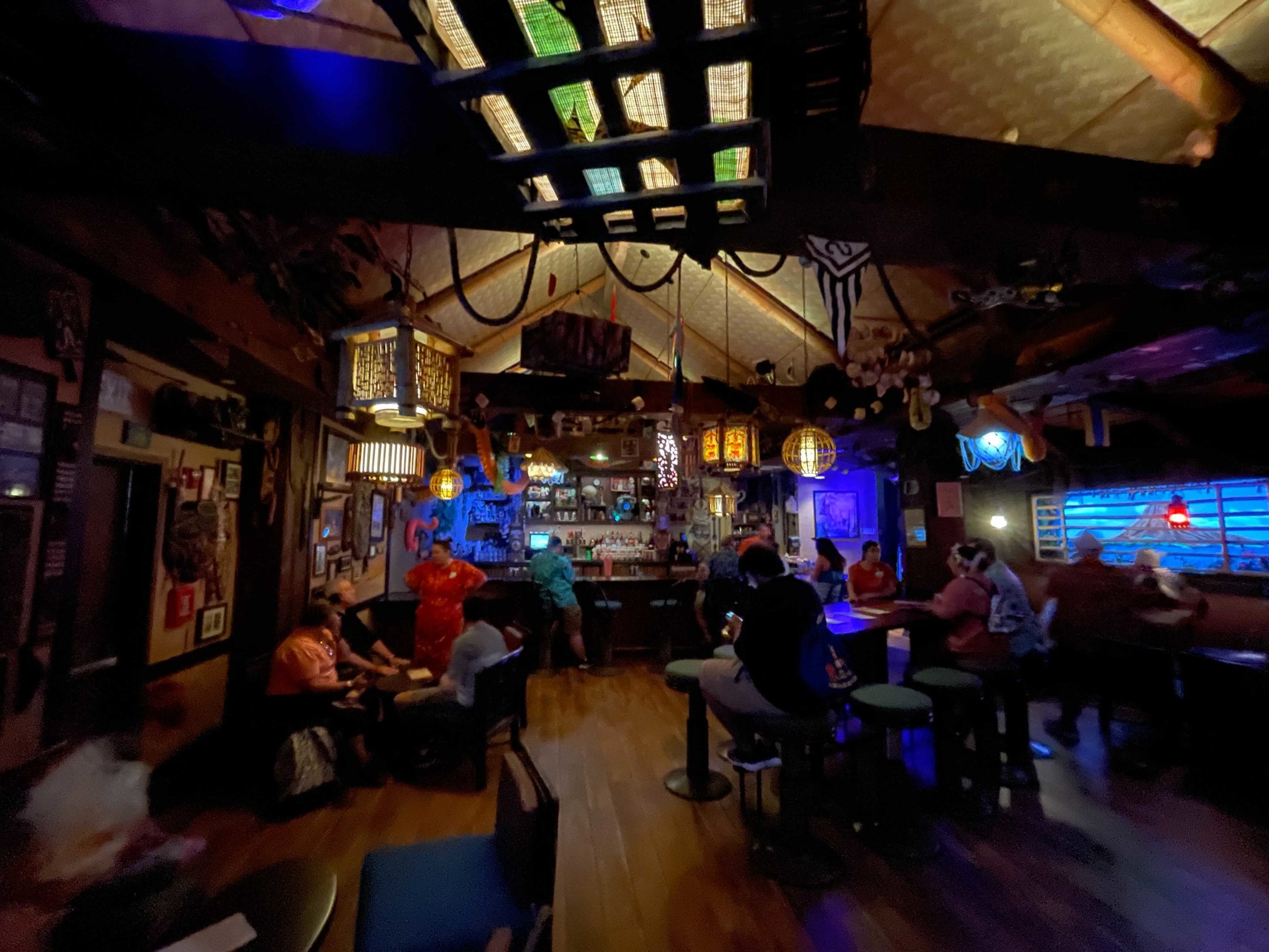 Dimly lit tiki bar with guests sipping cocktails beneath colorful lanterns, evoking Disney World’s Trader Sam’s Grog Grotto.