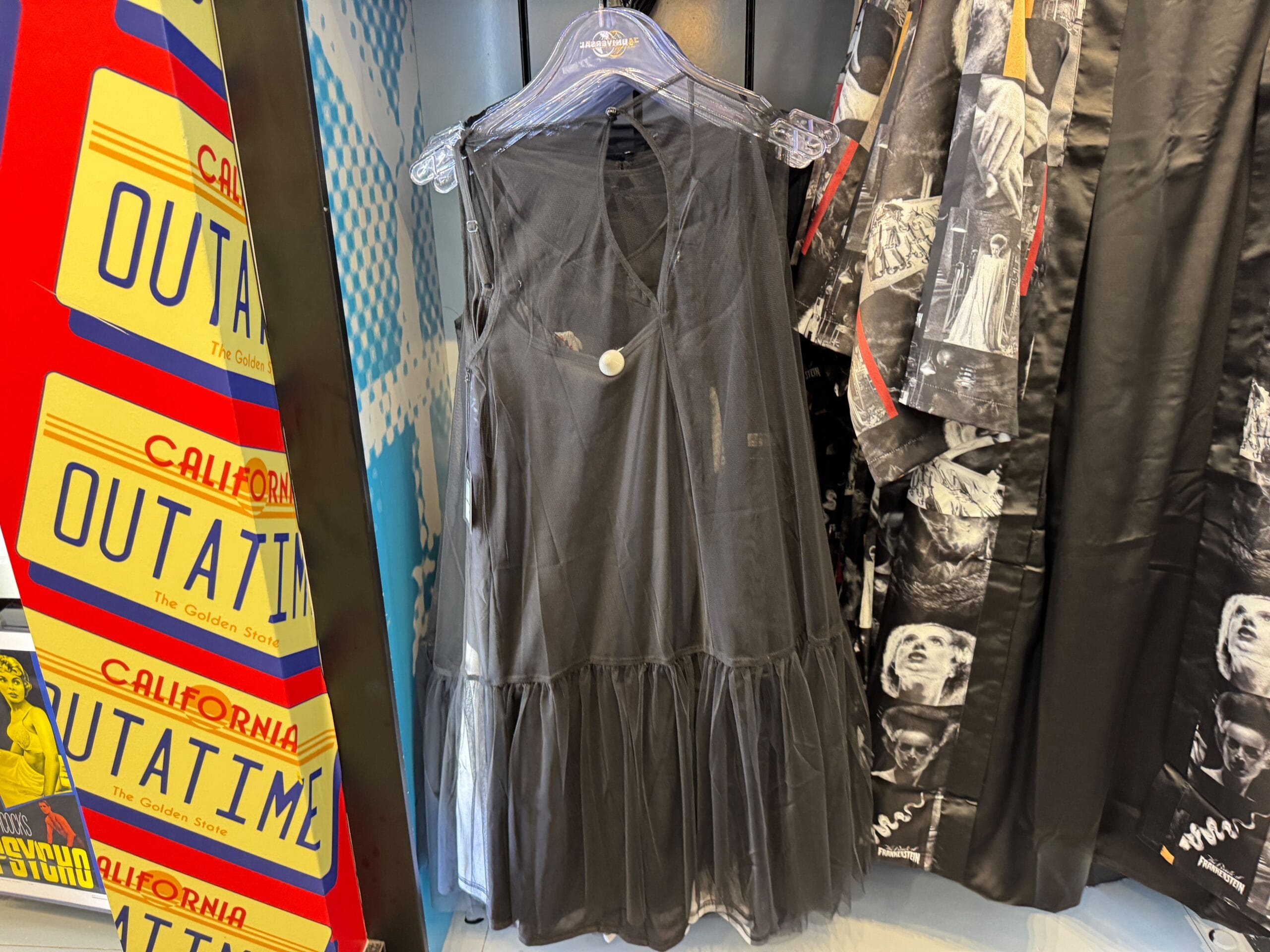 A sleeveless black dress with a sheer skirt hangs on a rack by clothes and California license plate signs, much like shops in Disneyland.