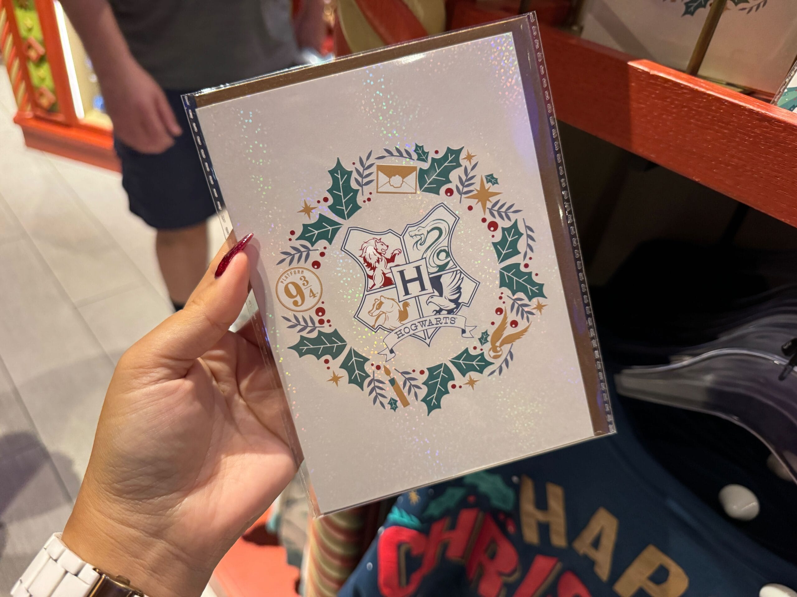 Hand holding a sparkly Harry Potter Christmas card with Hogwarts crest amid festive Hogwarts Merch in a store.