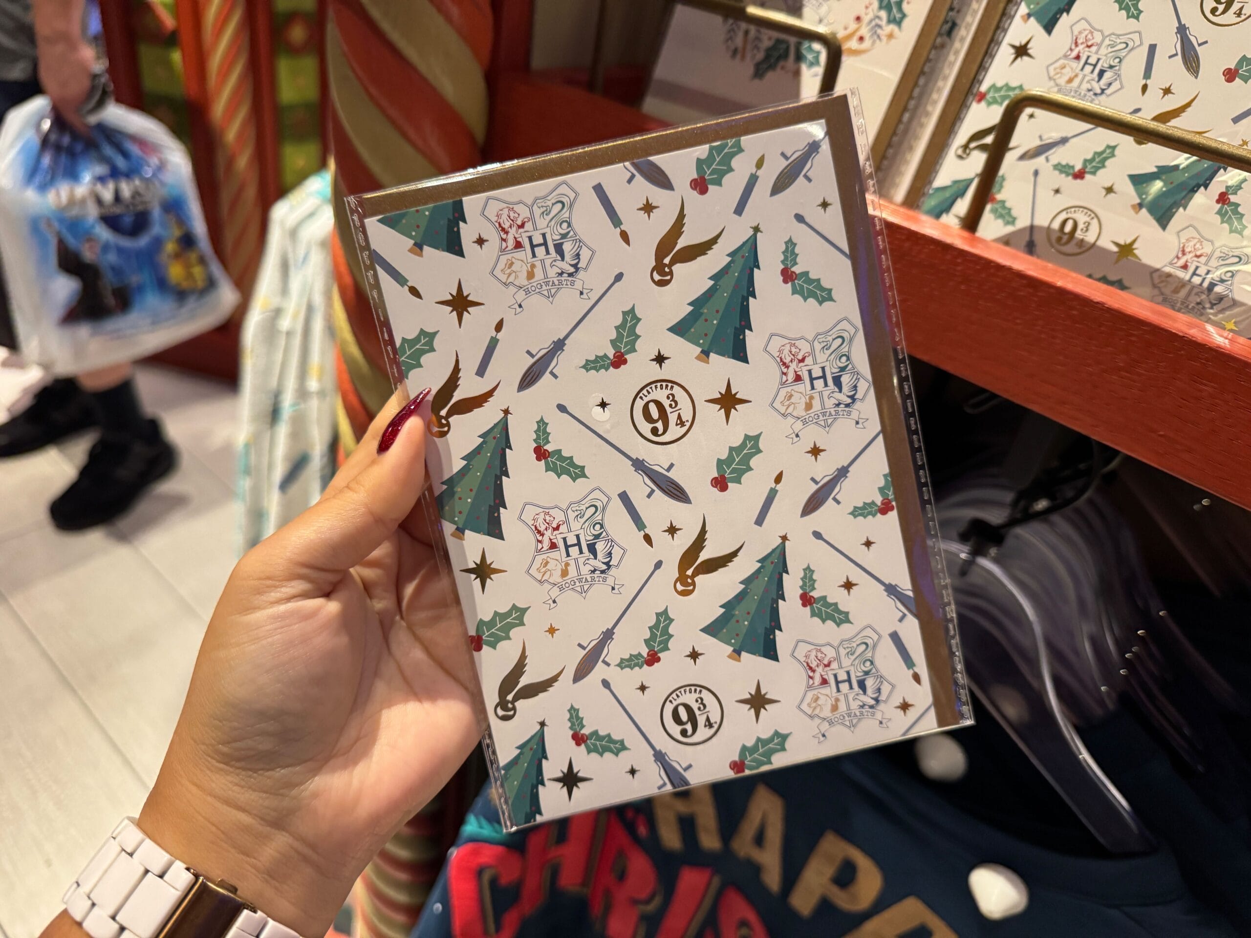 A hand holds a Hogwarts Merch notebook with icons like brooms, trees, wands, and Platform 9¾ on the cover.