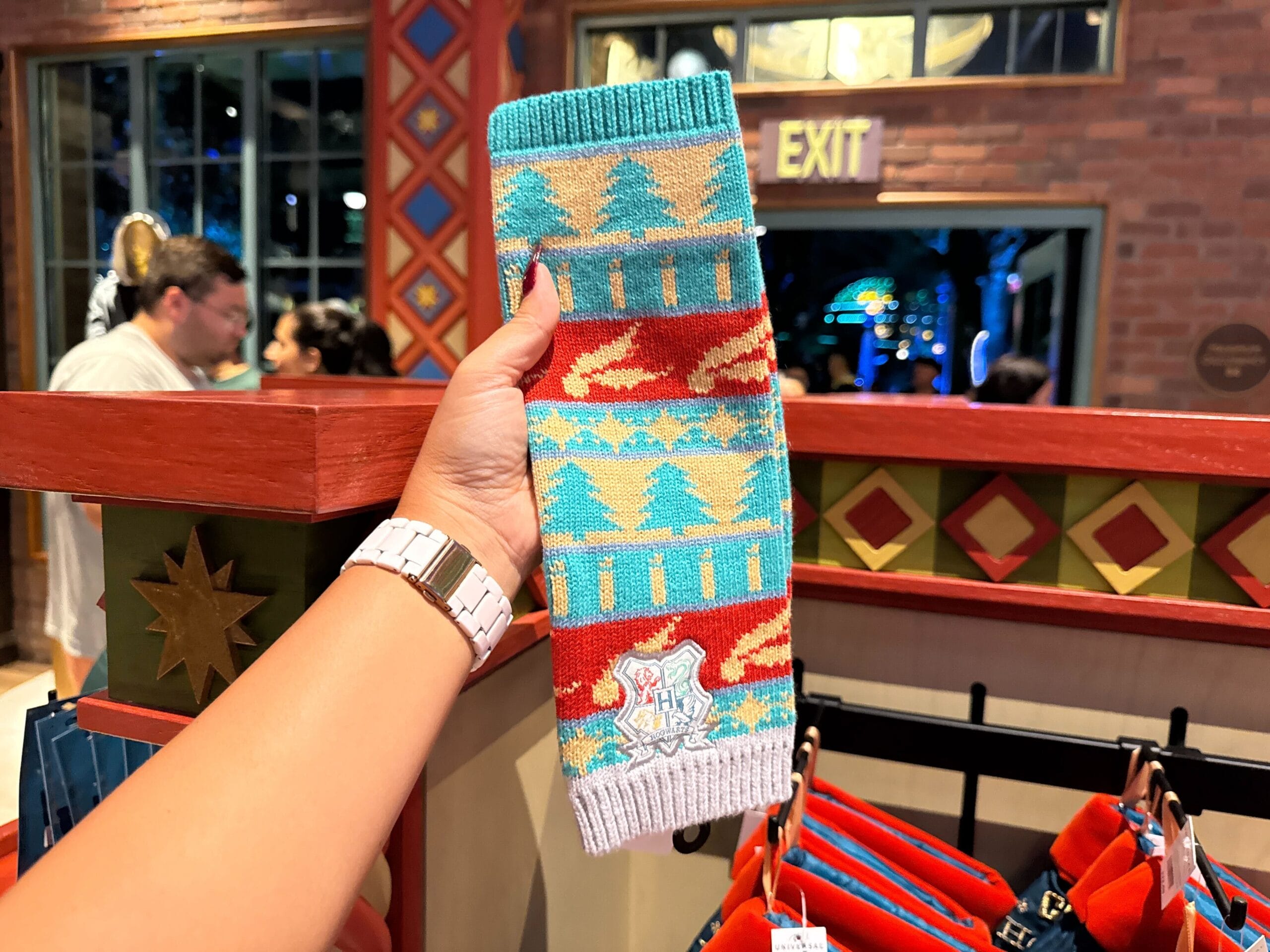 A hand holds a colorful Harry Potter Christmas knit scarf with trees and geometric patterns inside a store.
