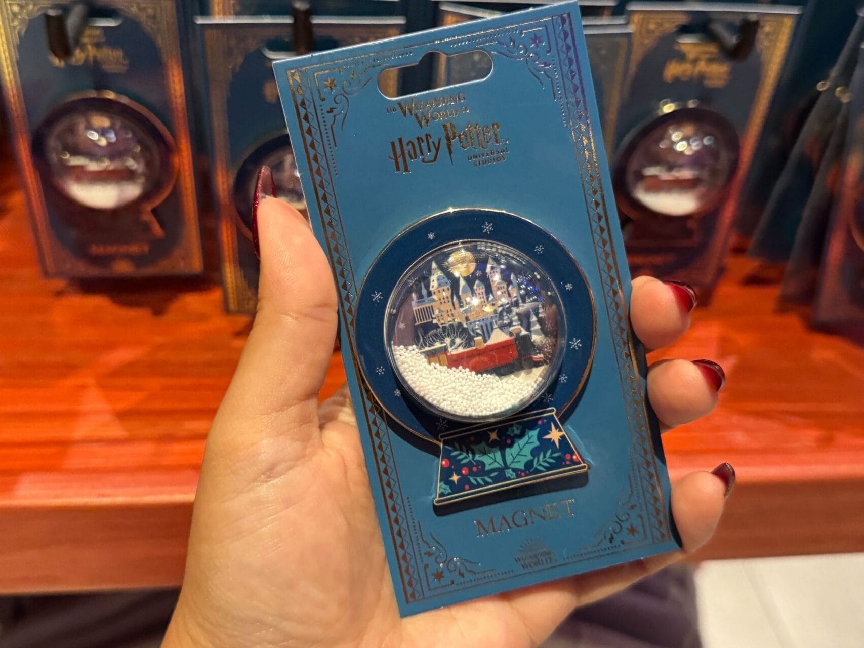 A hand holds a Hogwarts Merch magnet with several more displayed on a wooden shelf in the background.