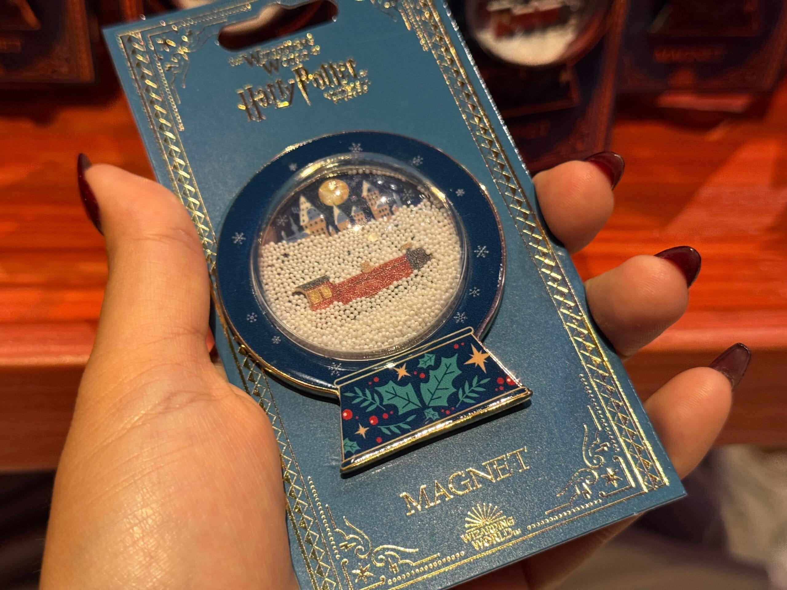 A hand holds a Harry Potter Christmas magnet featuring the Hogwarts Express in a festive snow globe design.