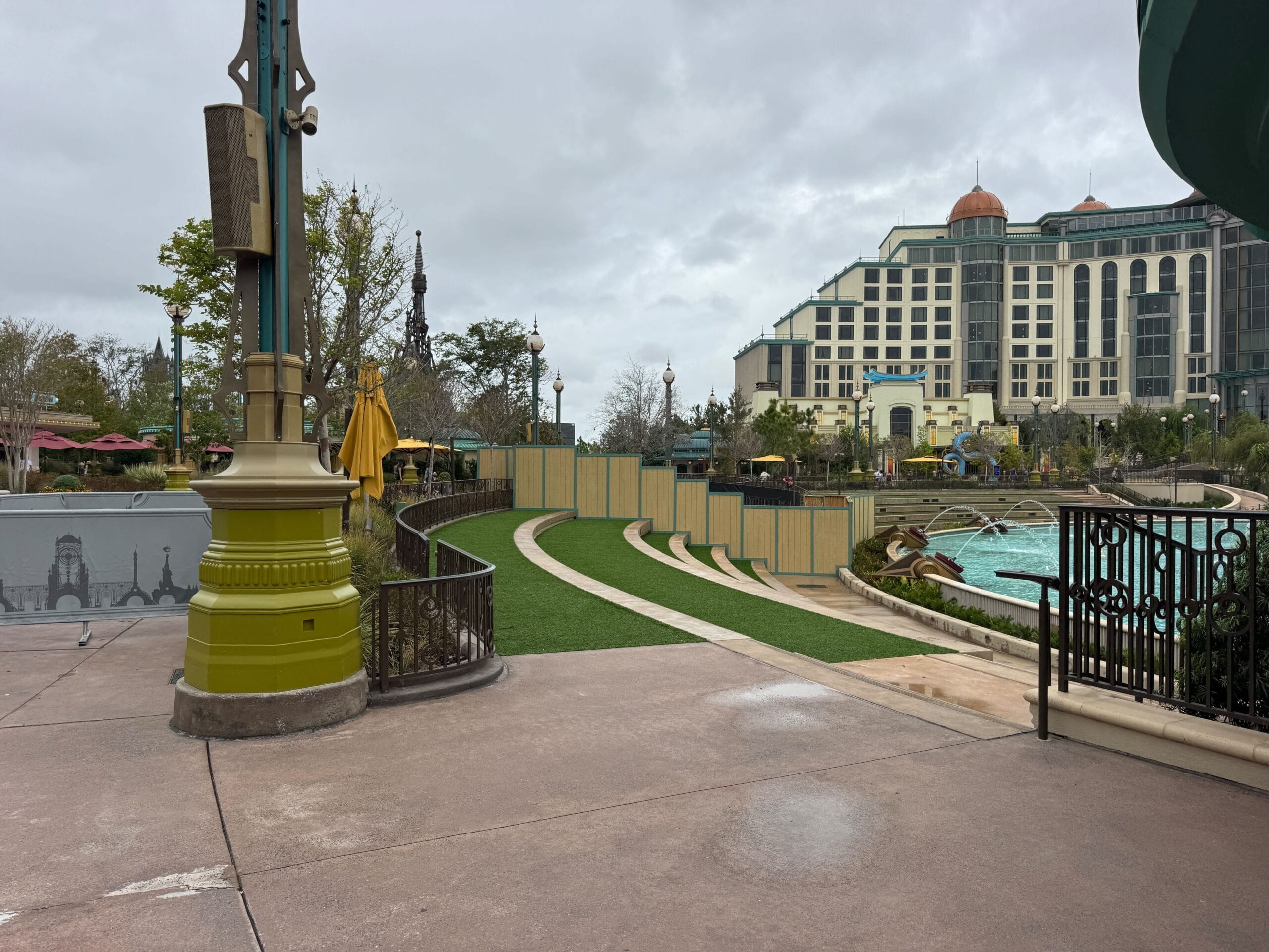 A curved walkway by a fenced lawn near a pool, with the Epic Universe hotel in the background.