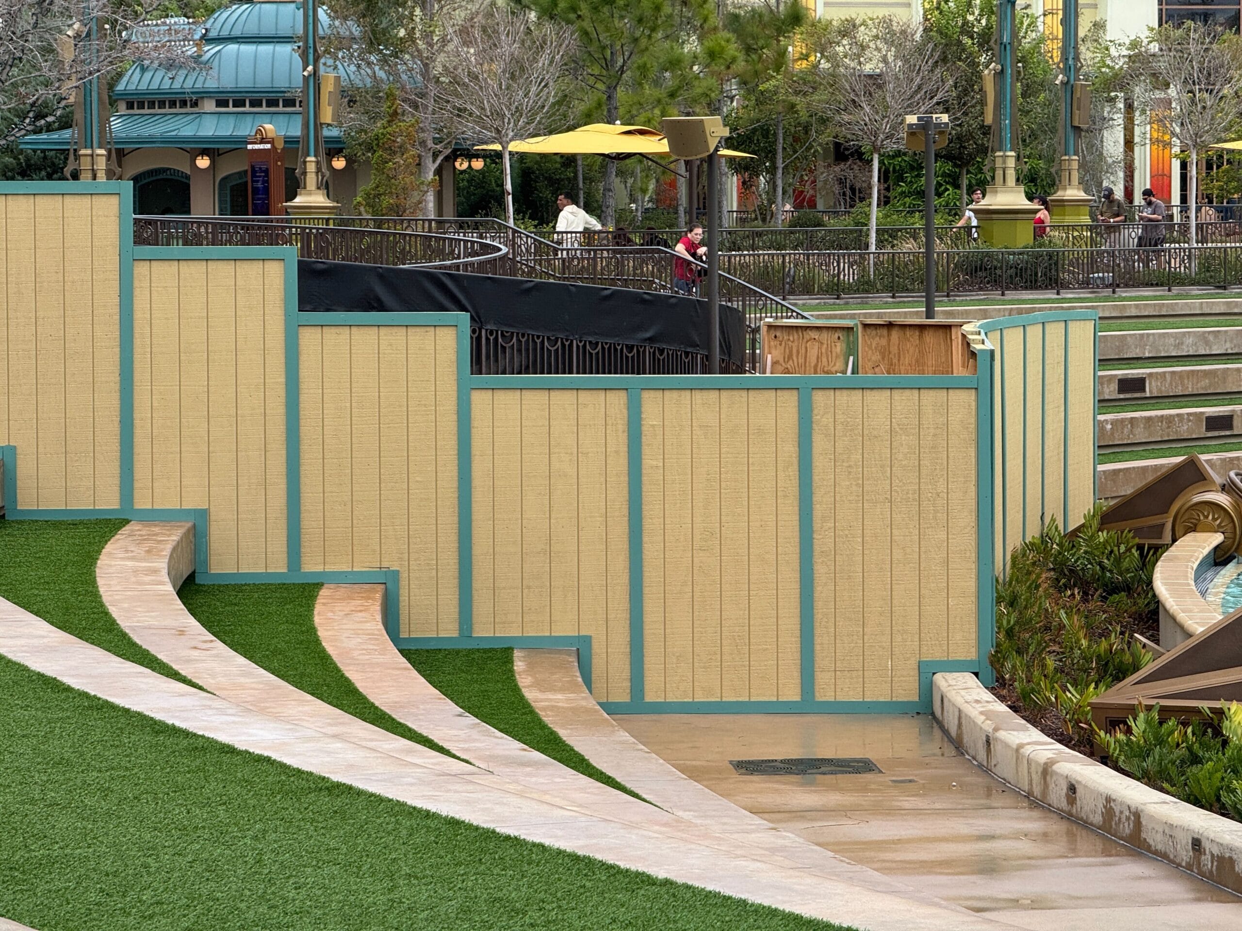 Construction walls block off a section of the park near the Mario Fountain, with artificial grass and curved concrete steps.