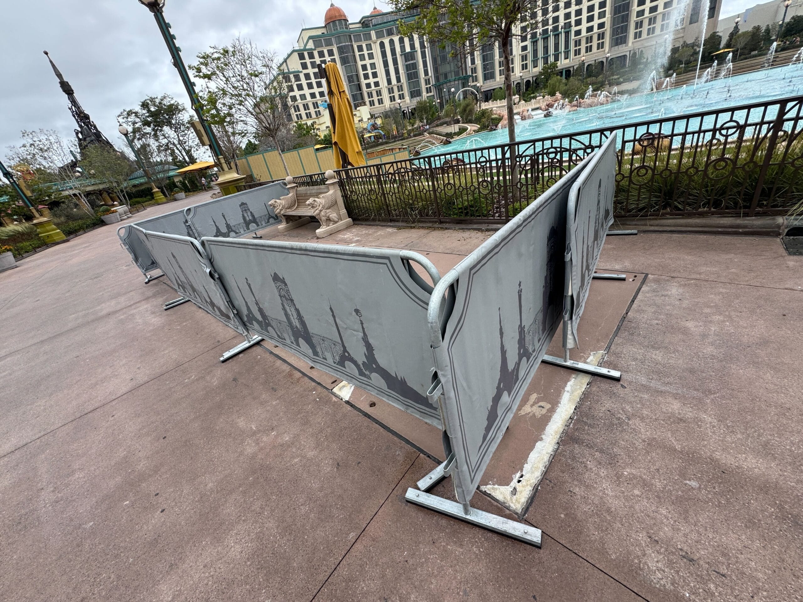 Grey construction walls block part of a walkway near a pool and hotel, with benches, trees, and the Mario Fountain nearby.