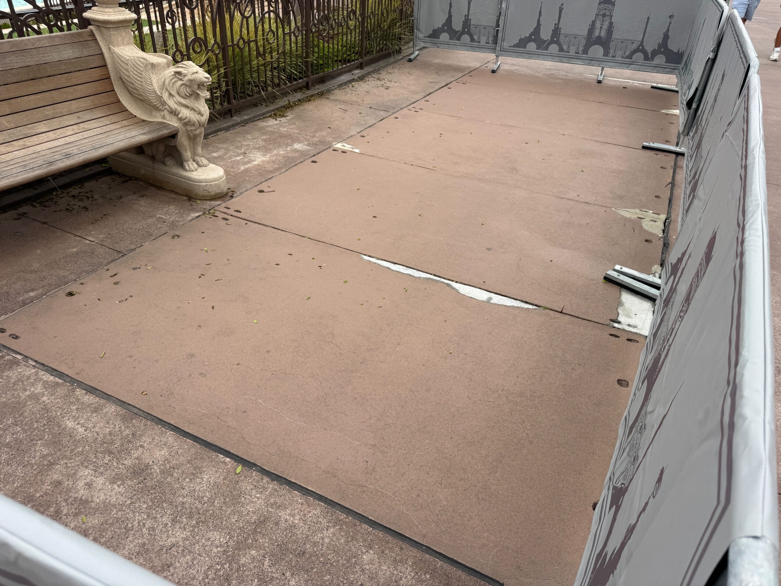 A sectioned-off area with tan pavement, a lion bench, and fencing with Eiffel Tower designs near the Mario Fountain.