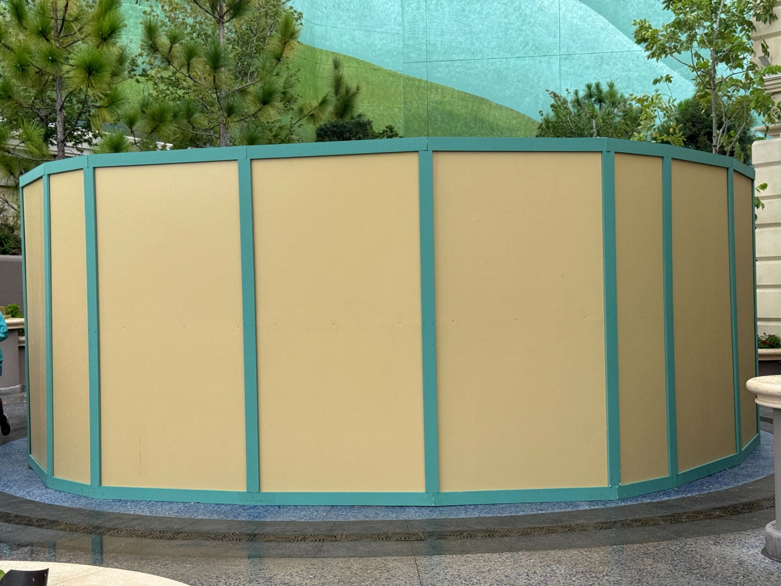 A curved beige construction wall with teal trim stands near trees at Epic Universe on a blue-tiled surface.