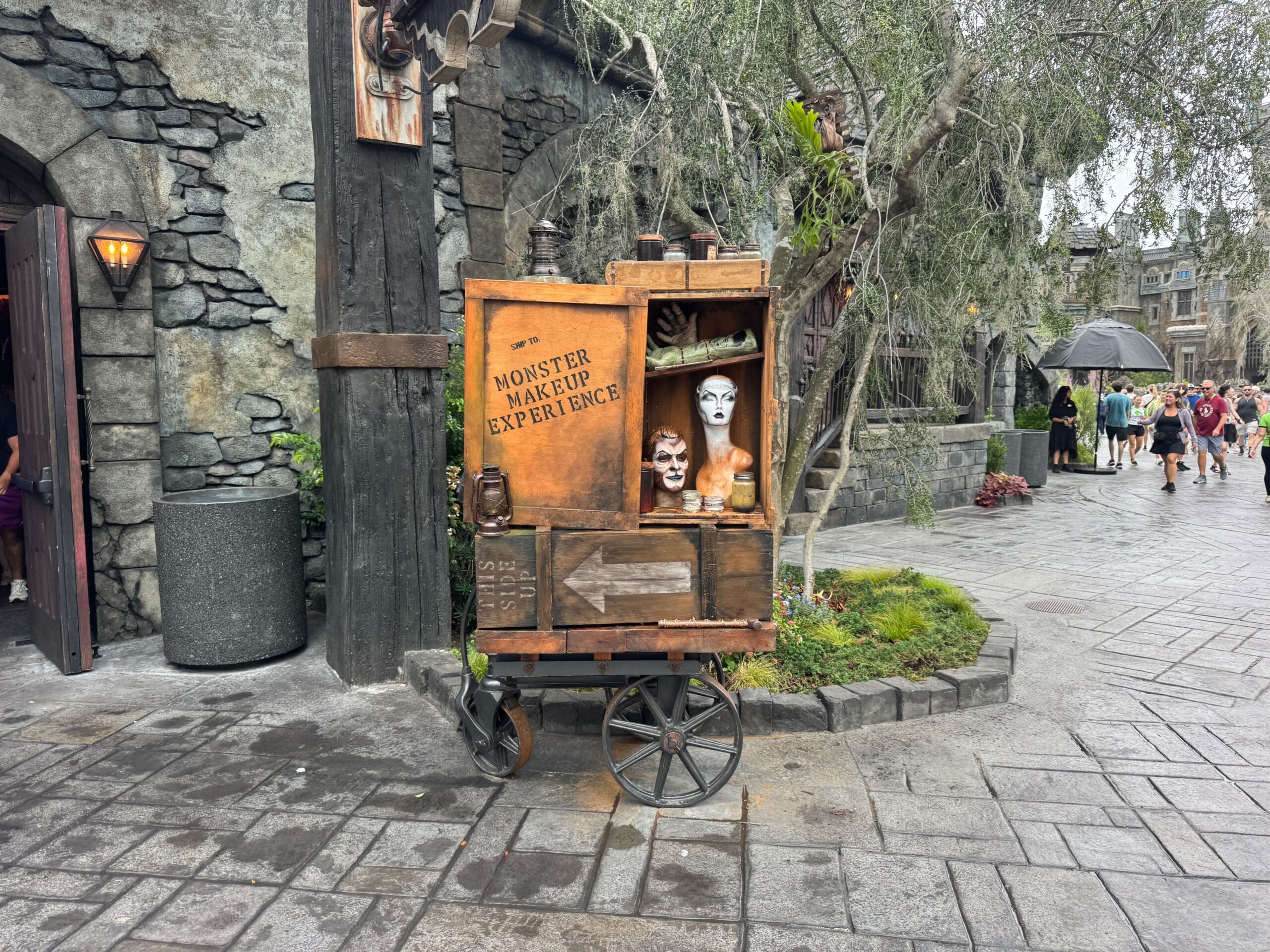 Epic Universe signage advertising showcases mannequin heads with monster makeup on a cart along the stone street.
