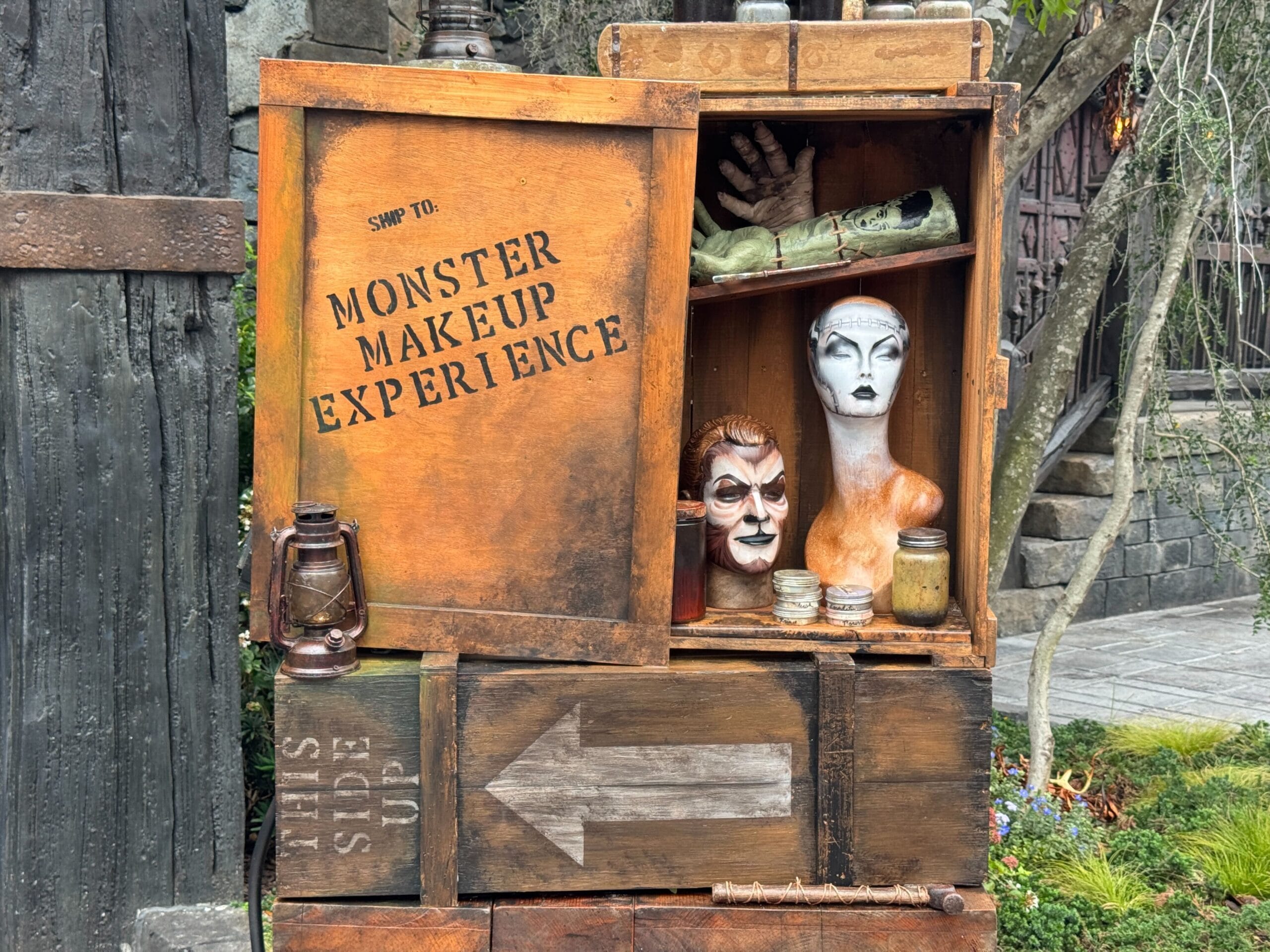 Wooden cabinet with Monster Makeup Experience signage, masks, props, and an arrow pointing left at Epic Universe.
