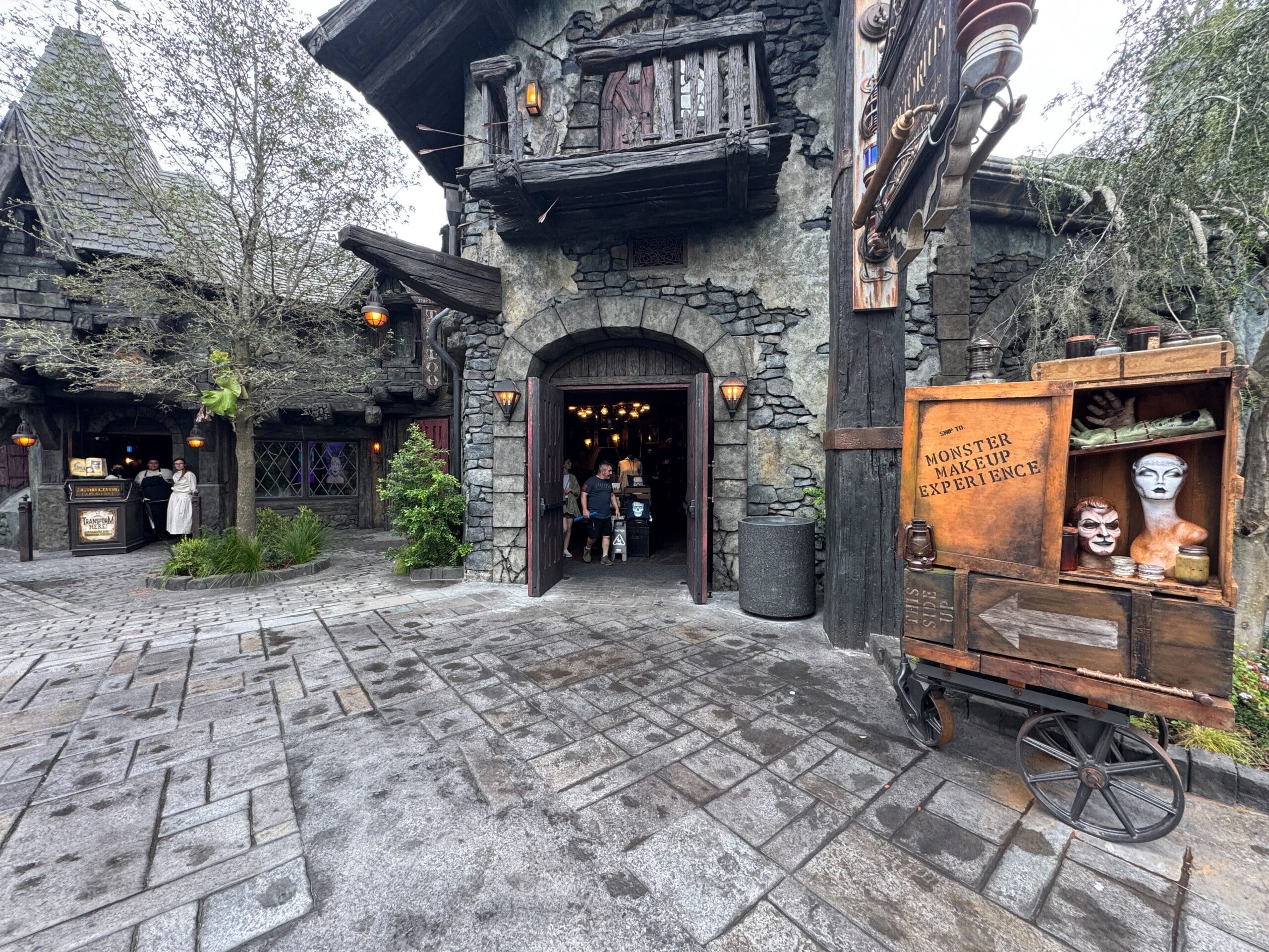 A stone building at Epic Universe with people entering, "Monster Makeup Experience" signage, and trees surrounding the area.
