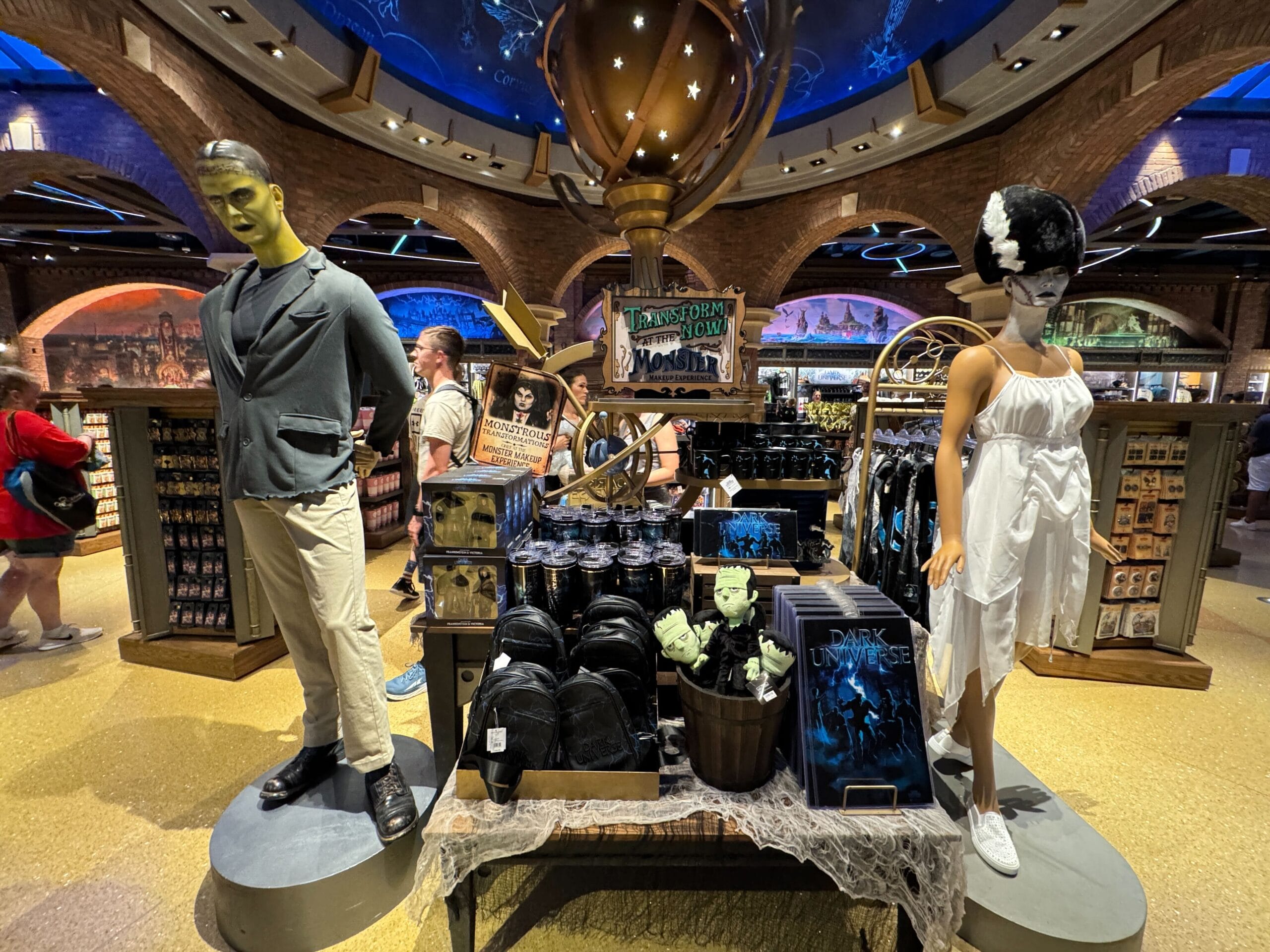 Store display with mannequins in themed costumes, Epic Universe backpacks, mugs, and Monster Makeup Experience merchandise.