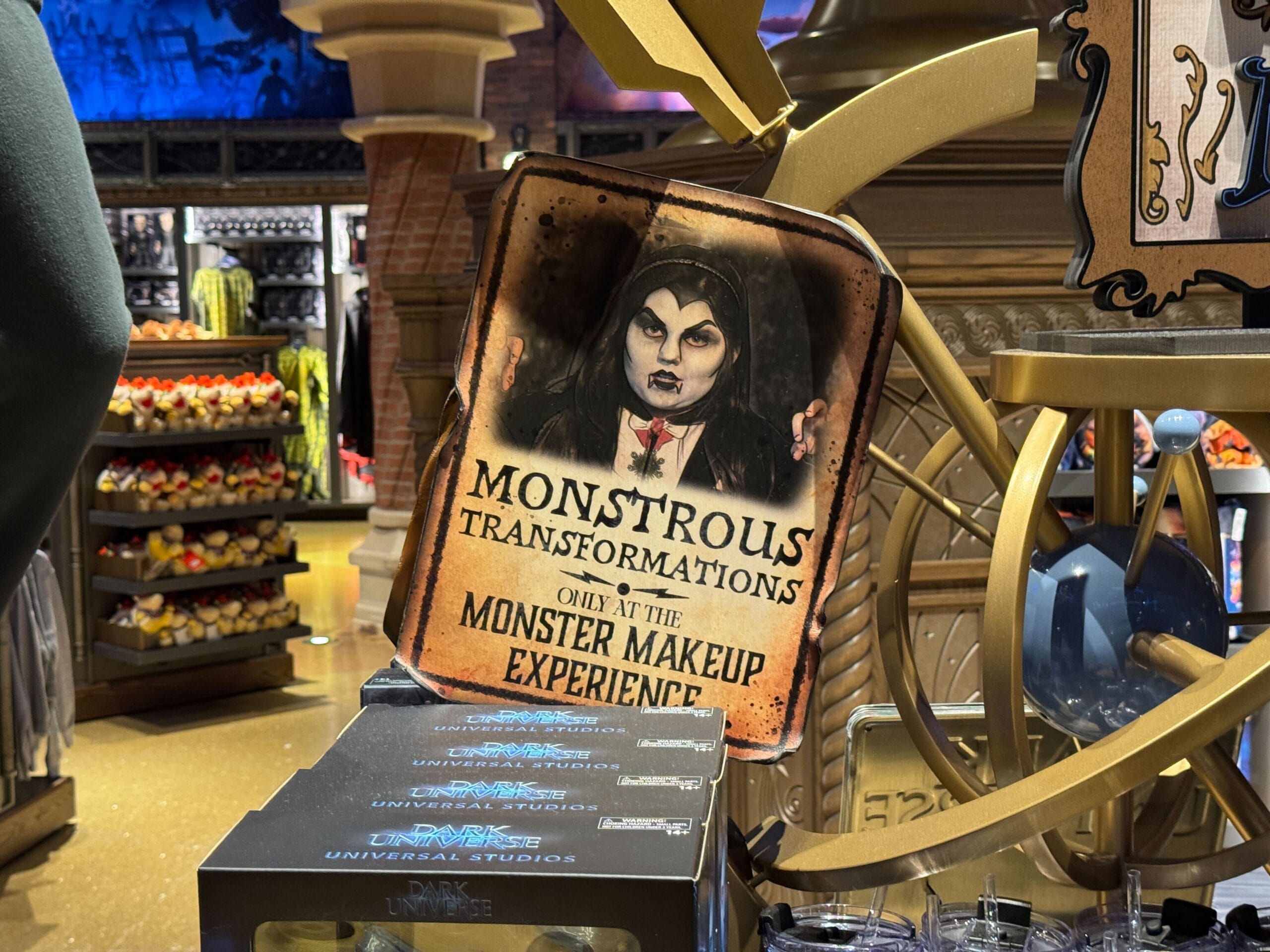 A sign for "Monstrous Transformations" with a vampire, promoting the Epic Universe Monster Makeup Experience.