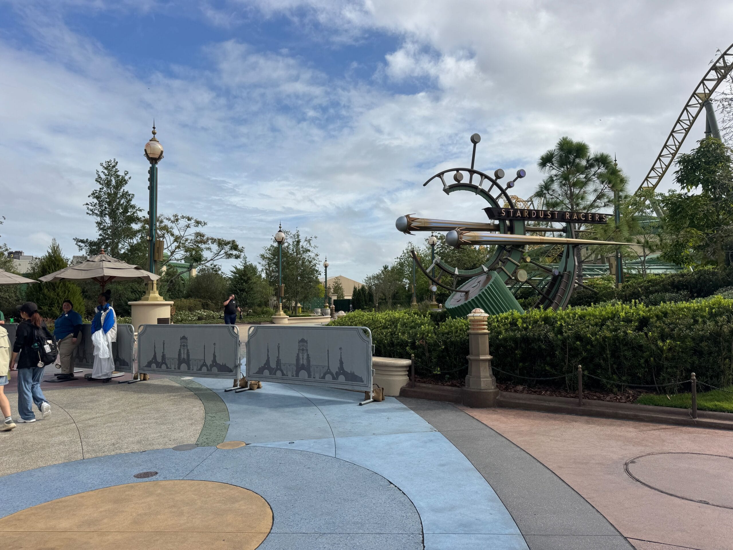 People stand near a construction barrier as Stardust Racers prepares for reopening at the theme park on a partly cloudy day.