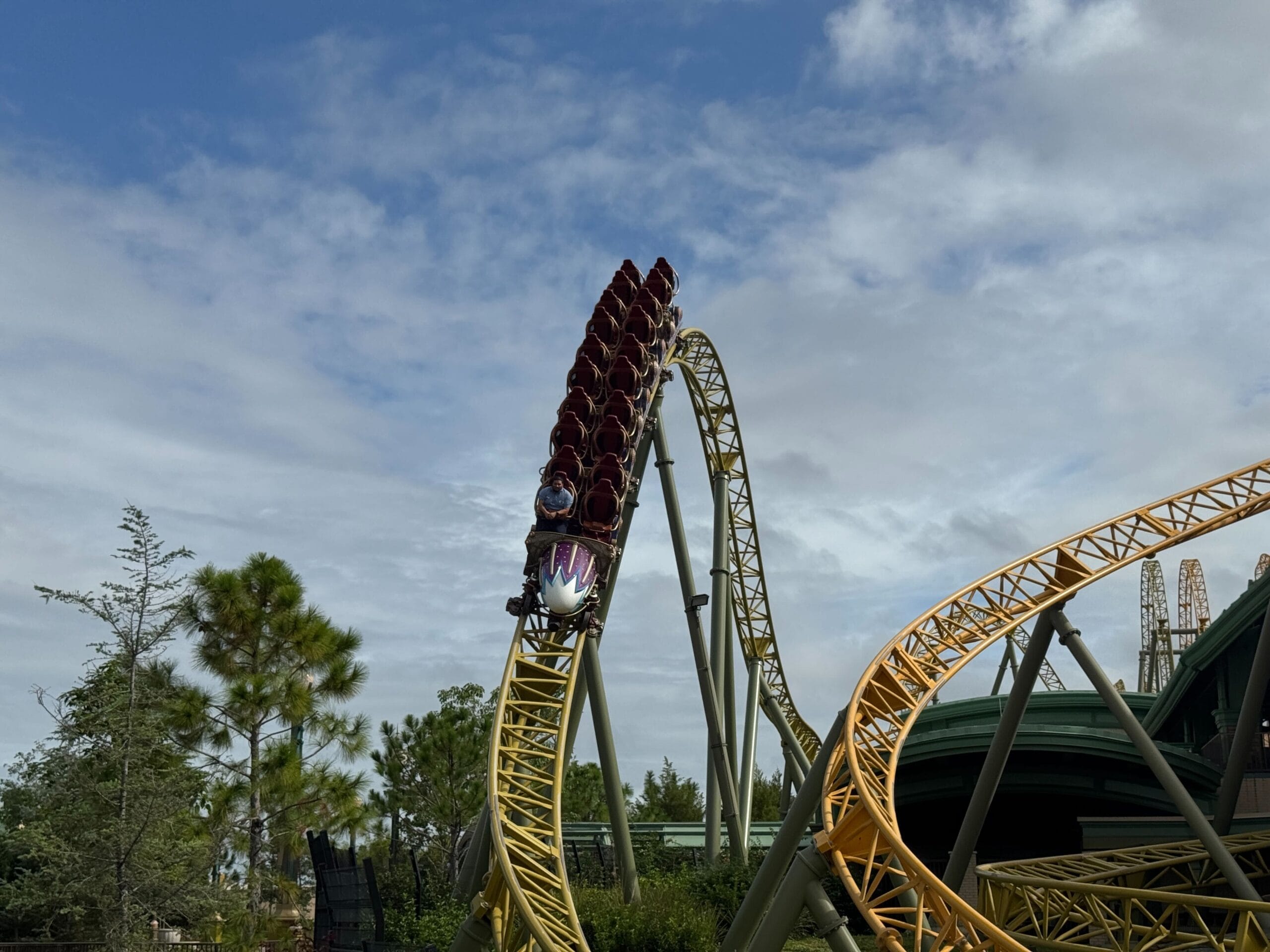 A roller coaster train speeds down the yellow Stardust Racers track with riders, testing under a partly cloudy sky.