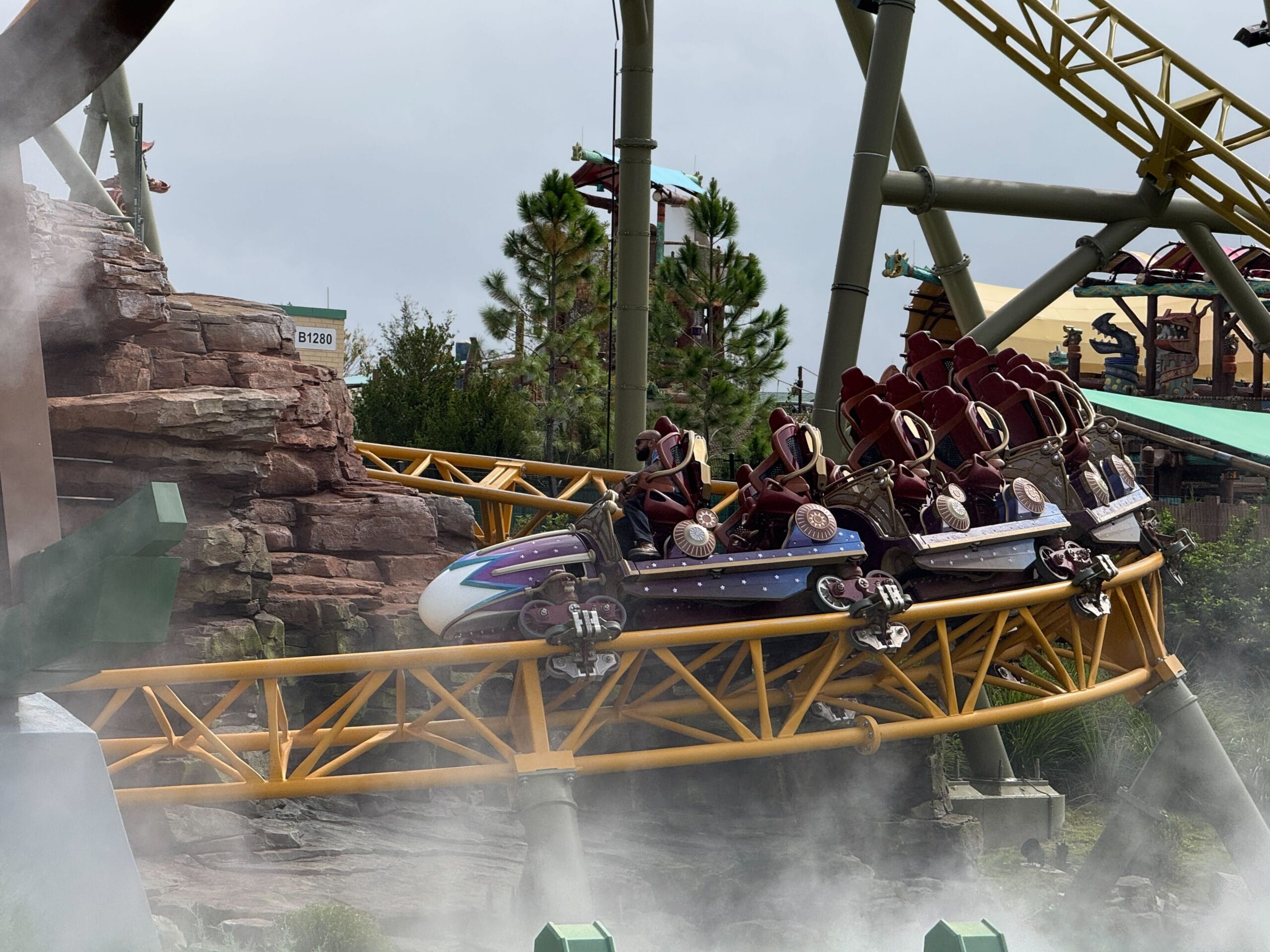 People testing the yellow Stardust Racers roller coaster, curving through rocks, mist, and greenery under a cloudy sky.