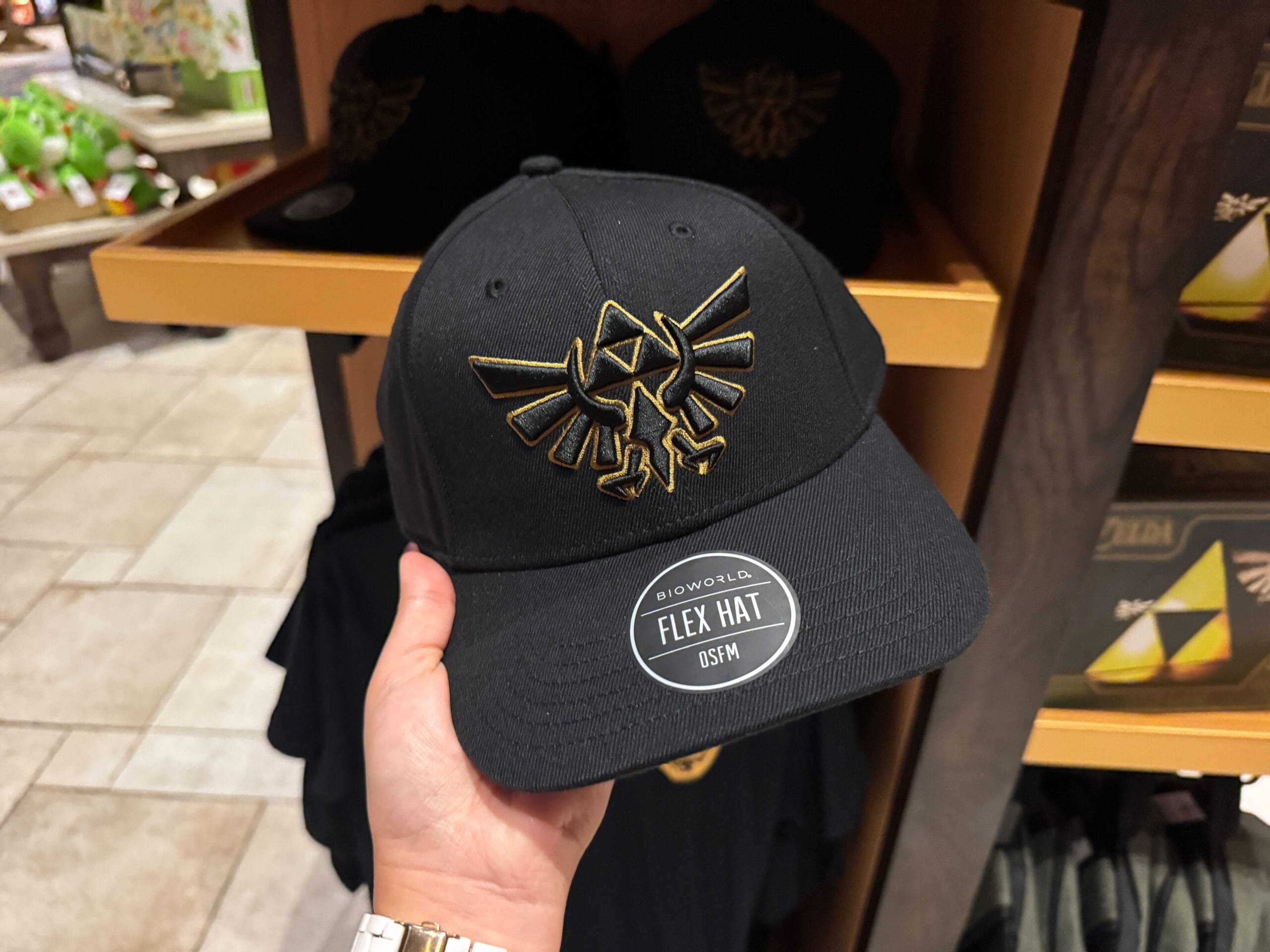 A hand holds a black Zelda merchandise hat featuring a gold Hylian crest and a "FLEX HAT" sticker on the brim.