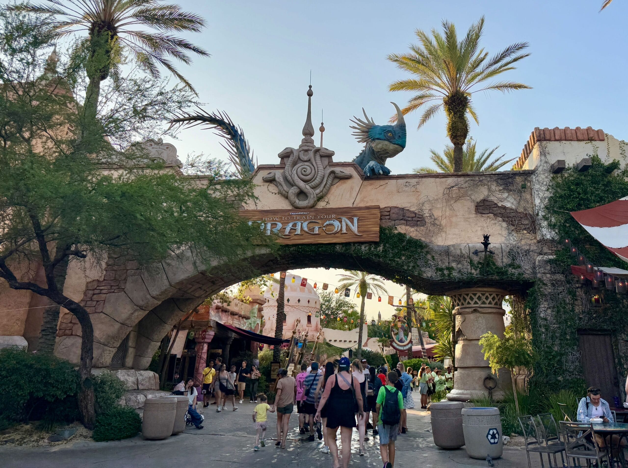 Guests stroll beneath a stone archway topped with a dragon and "DRAGON" sign at a theme park, unlike any Disney Castle.