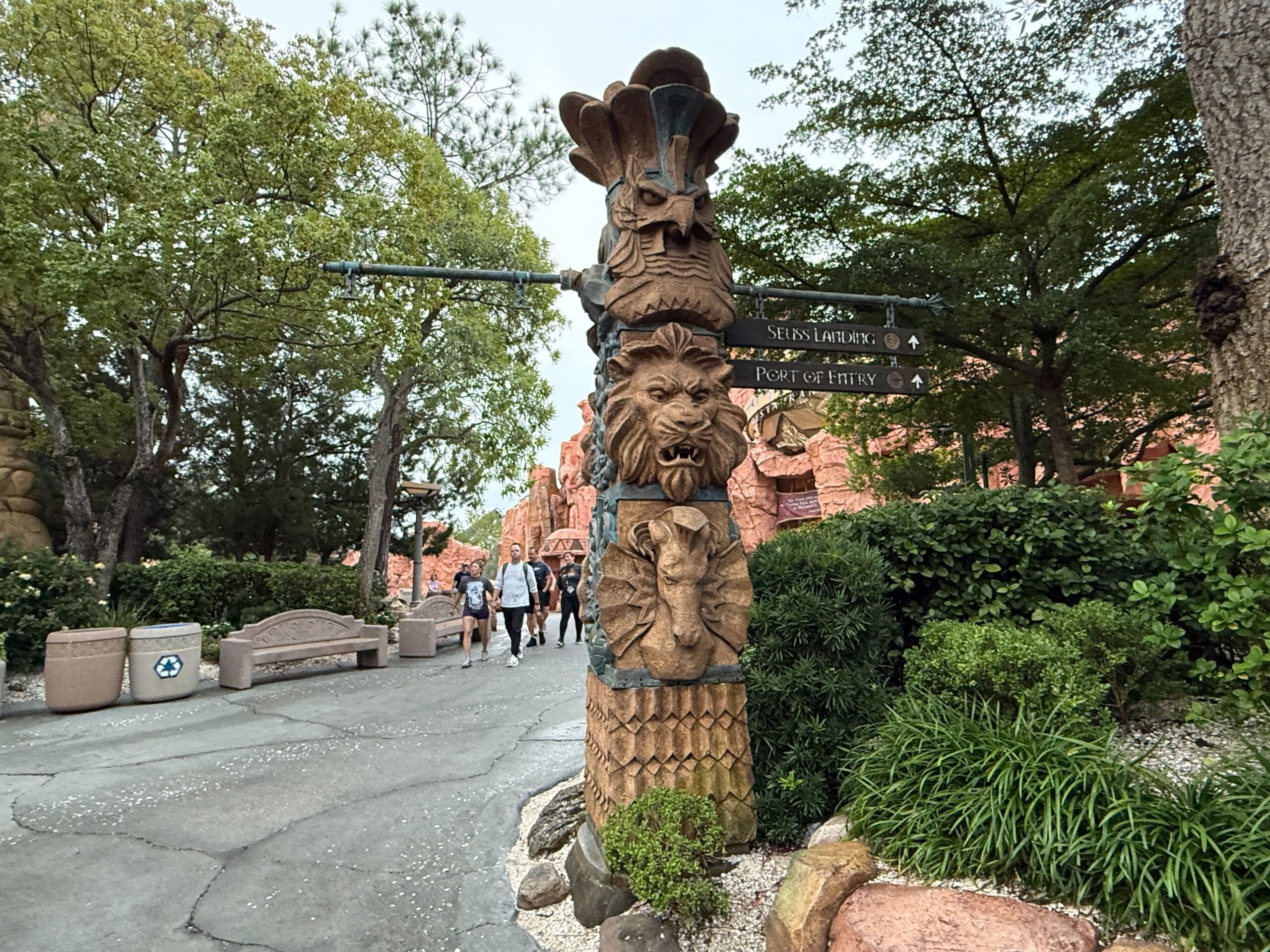 A carved totem pole with animal faces stands by directional signs on a lush path, evoking Adventureland in Disney parks.