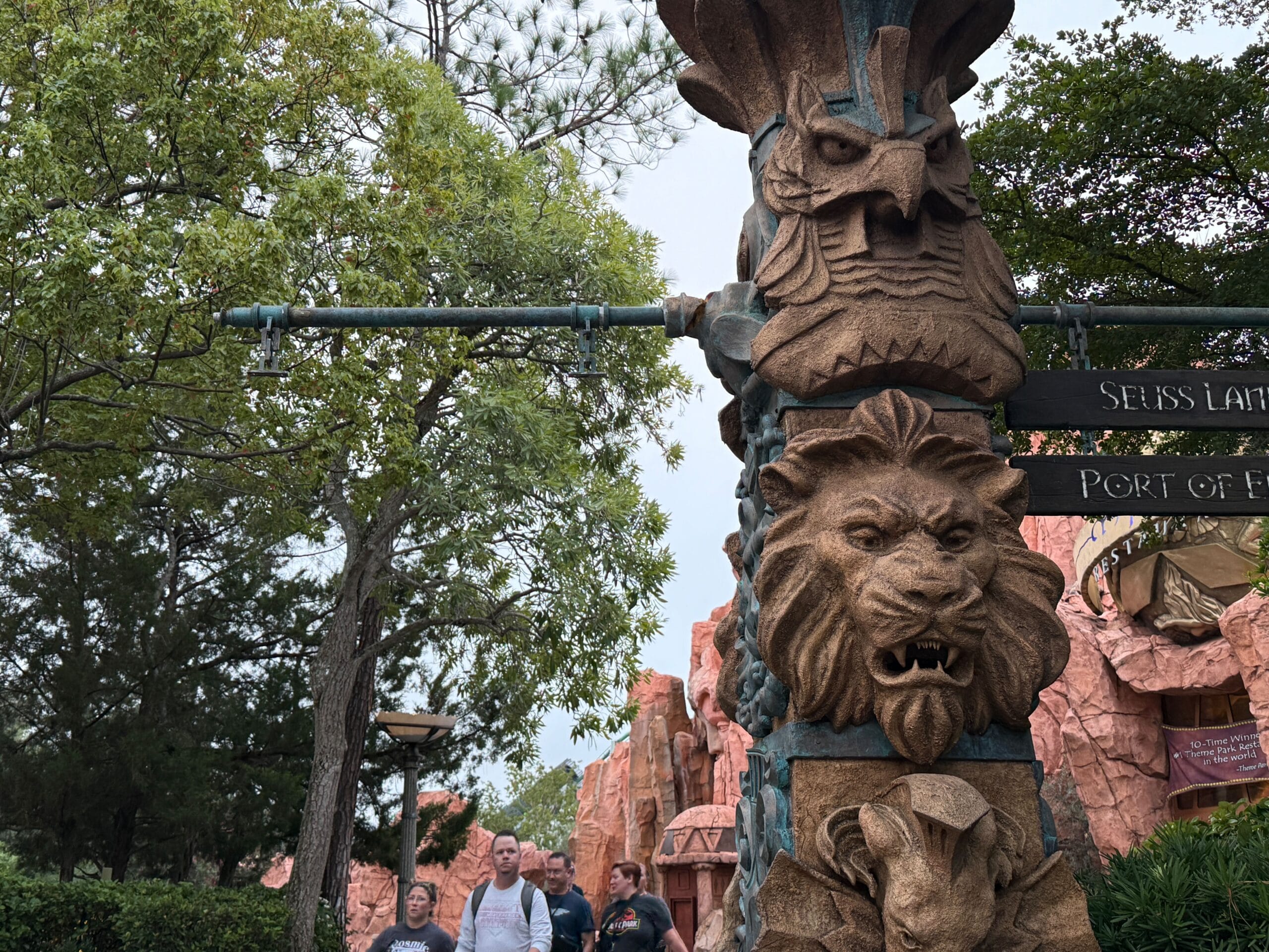 A themed stone pillar with animal carvings and directional signs, reminiscent of Disney parks; guests stroll amid lush landscaping.