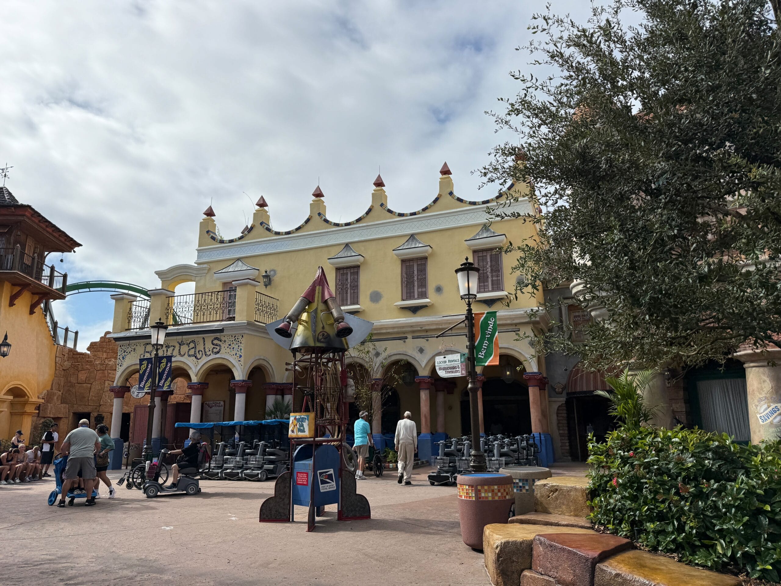 Guests stroll by a vibrant, Mediterranean-inspired building with outdoor seating and towers, evoking Disney park architecture.