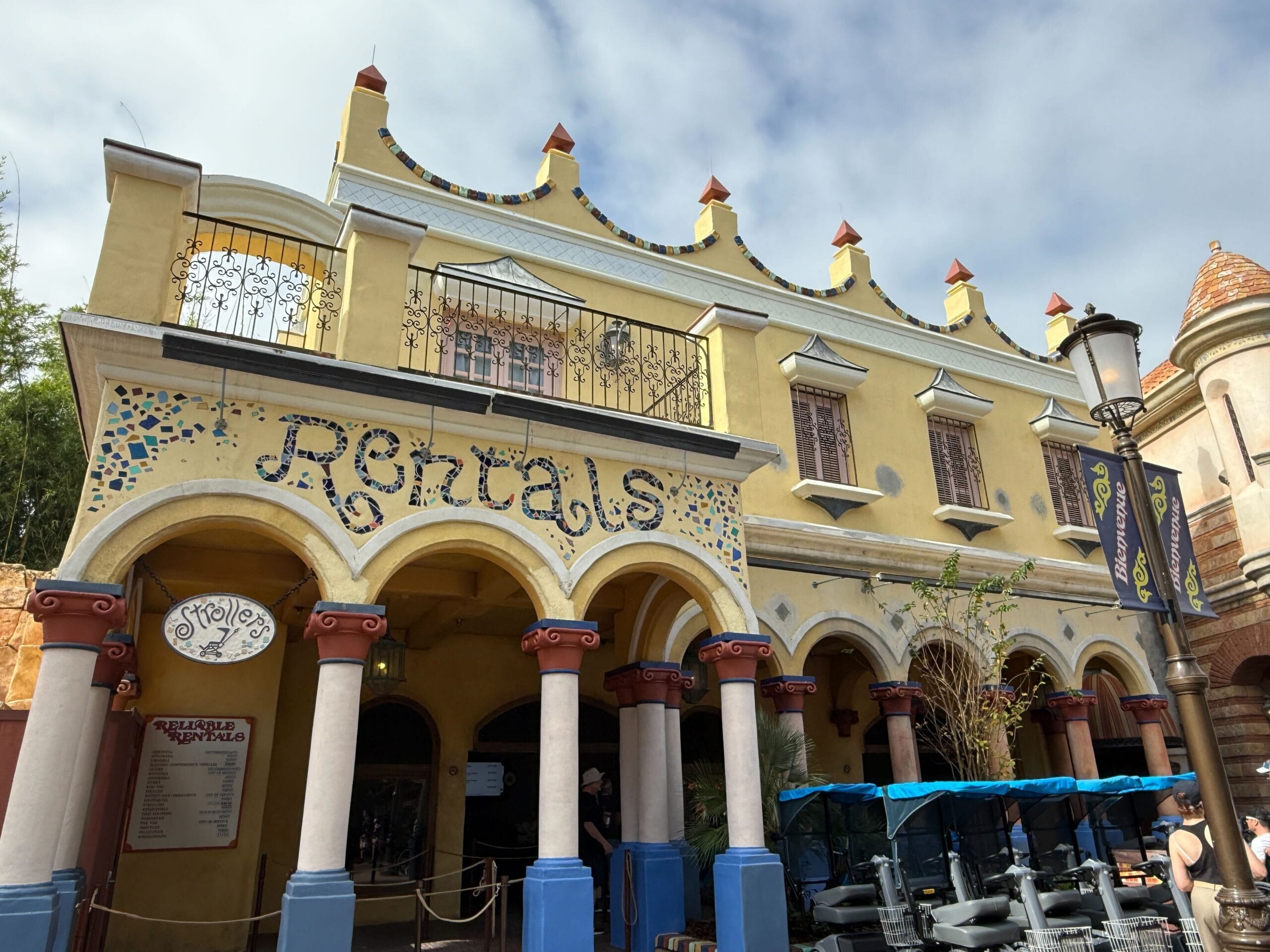 A yellow "Rentals" building with arched entryways and strollers outside, reminiscent of Main Street décor in Disney Parks, under clouds.