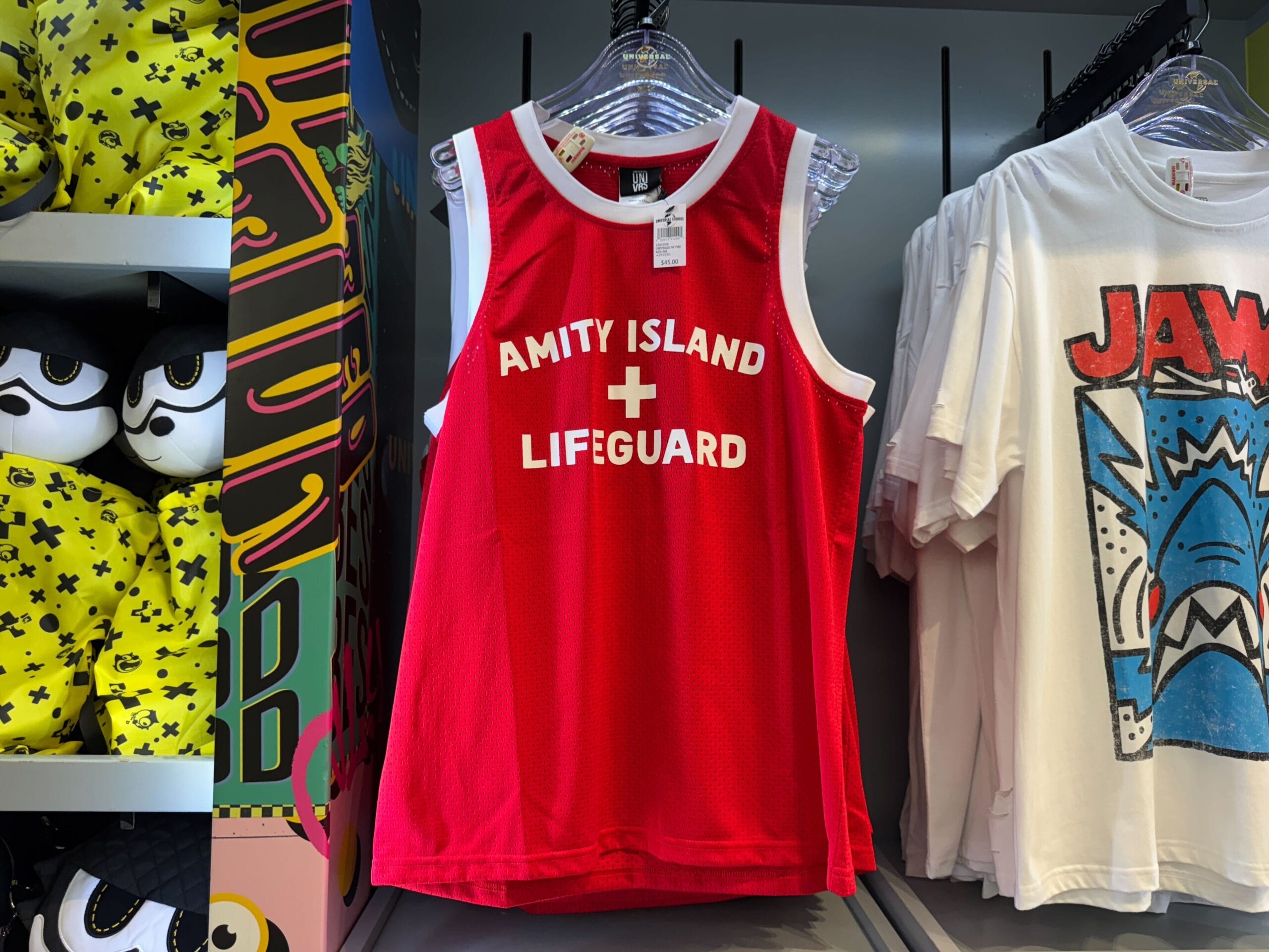 A red "Amity Island Lifeguard" tank top and white "Jaws" t-shirt hang on a store rack, reminiscent of Universal, not Disney.
