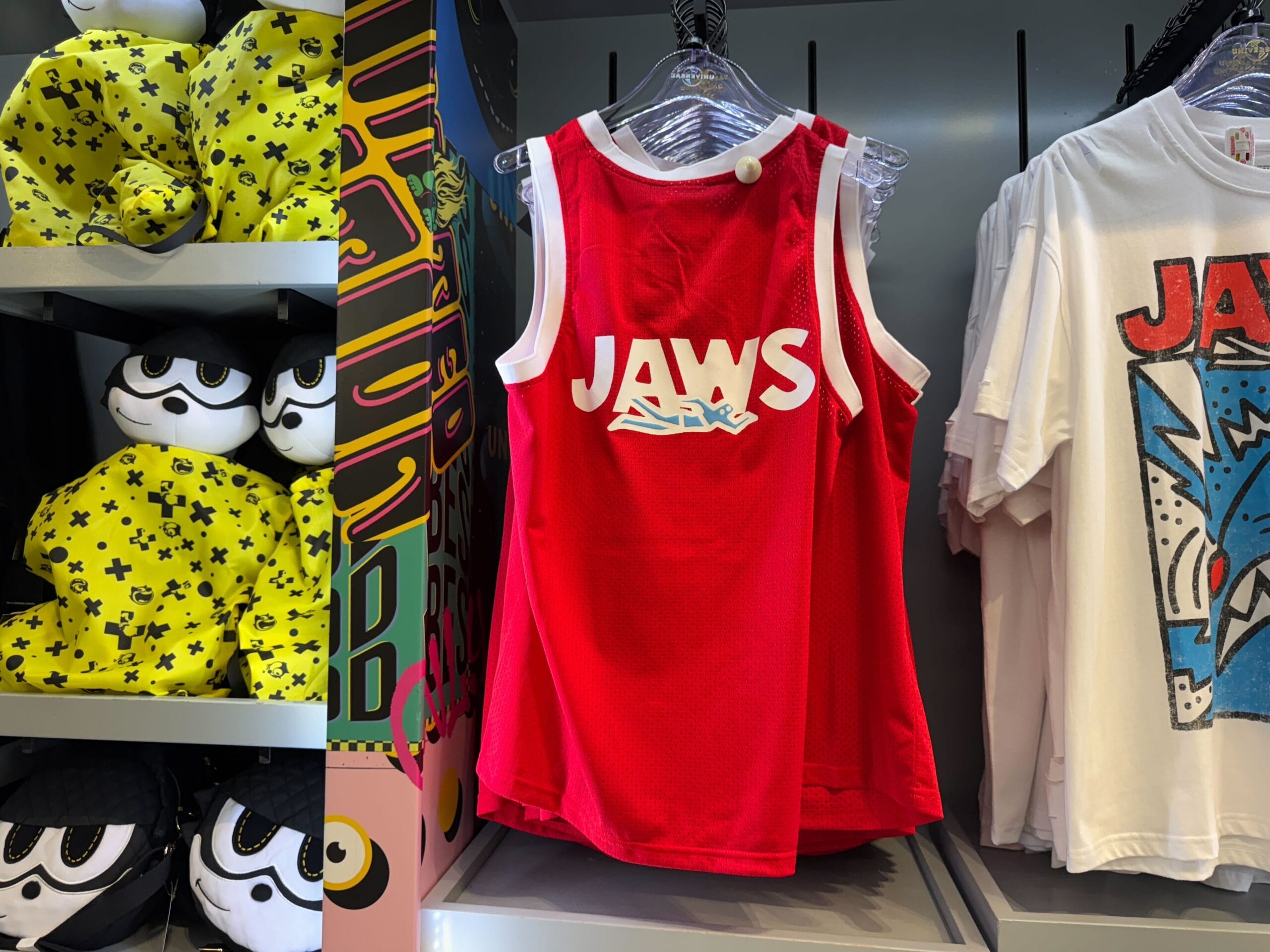 A red "JAWS" tank top hangs on a rack beside white Jaws shirts and yellow plush toys, echoing iconic theme park merch displays.