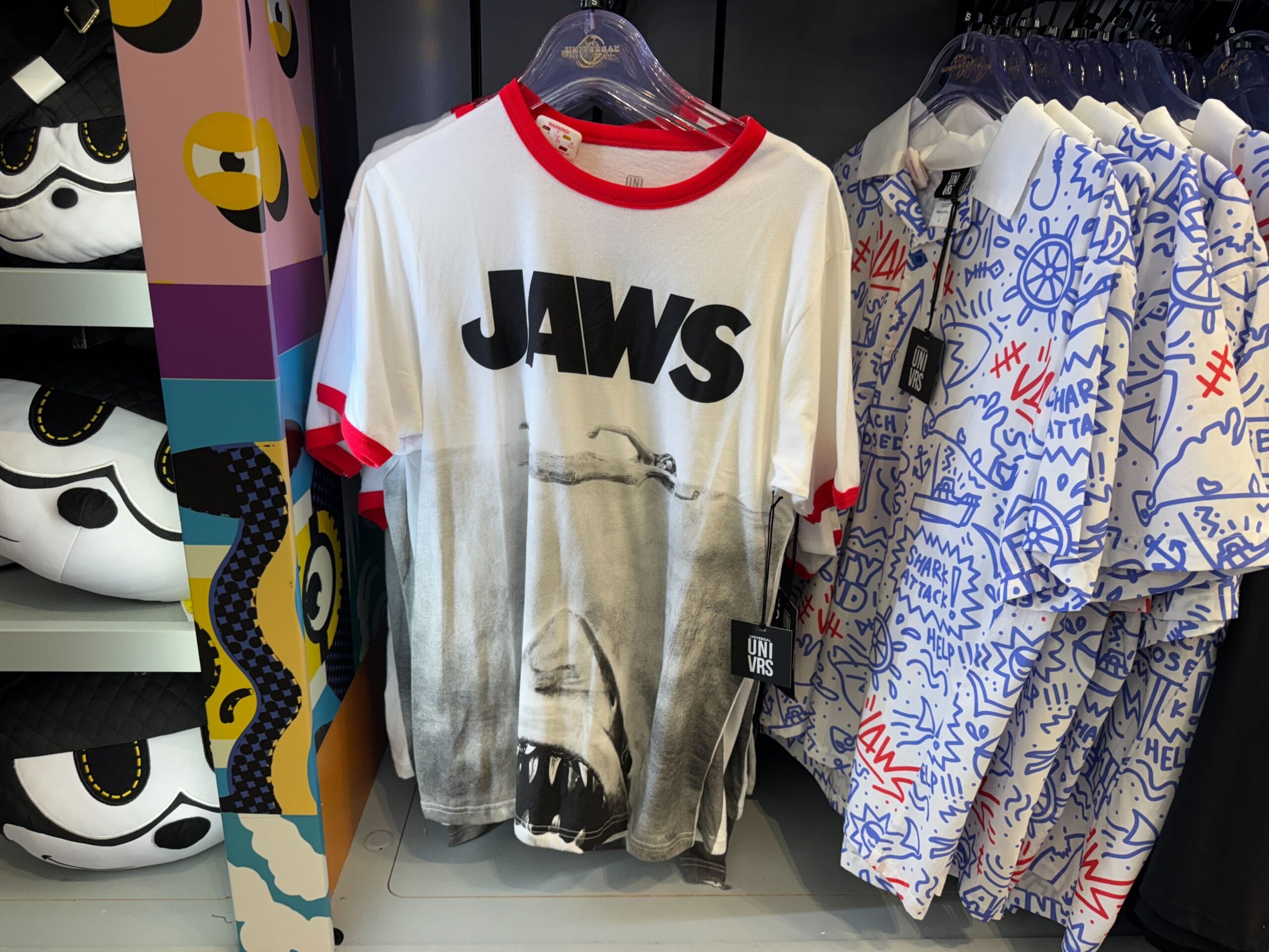 A white JAWS t-shirt with a shark graphic hangs among colorful shirts in a shop, reminiscent of Disney Parks’ themed stores.