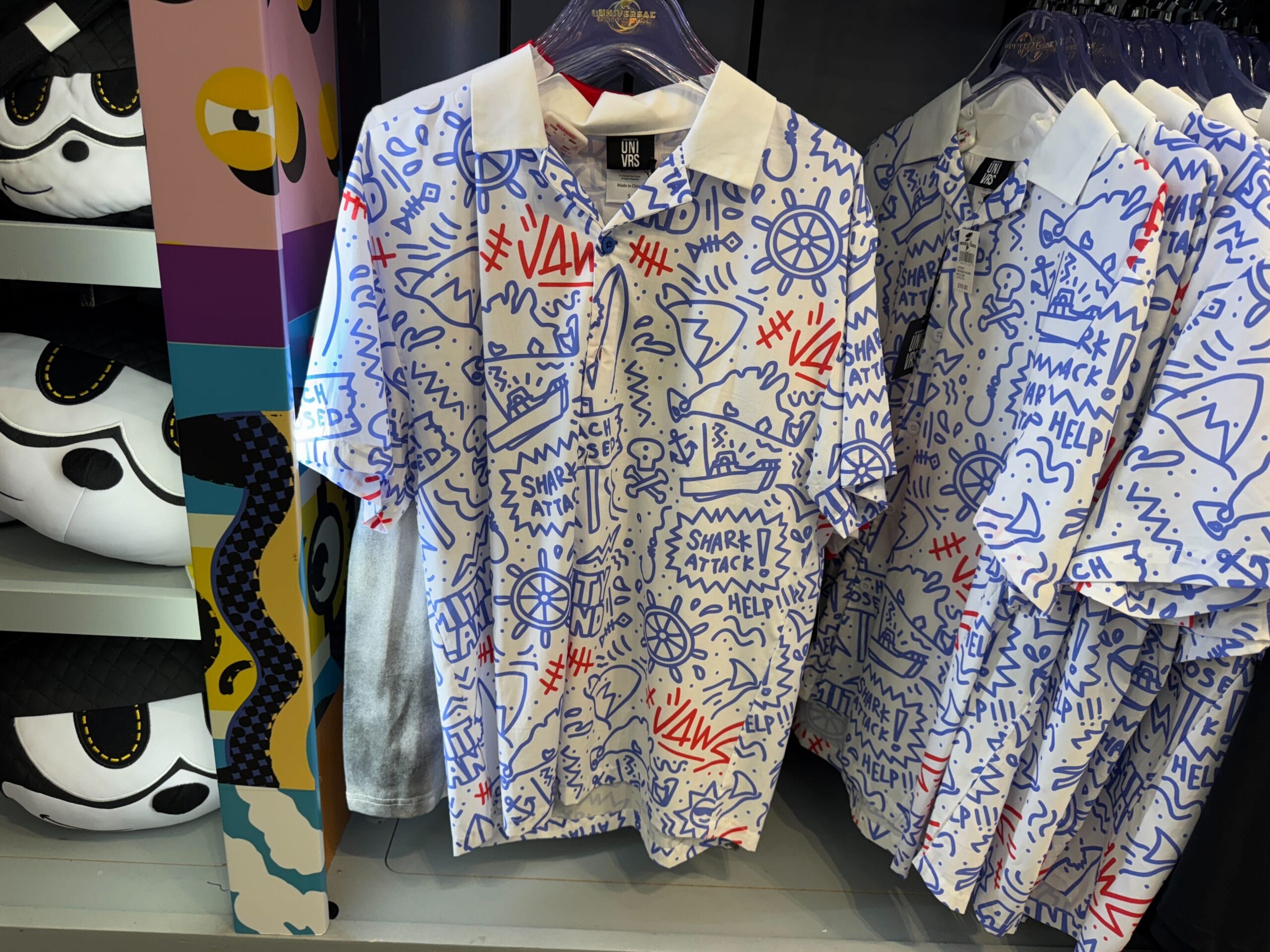 White polo shirts with playful blue and red doodles, such as "SHARK ATTACK" and "HELP," hang on racks, not Disney-branded.