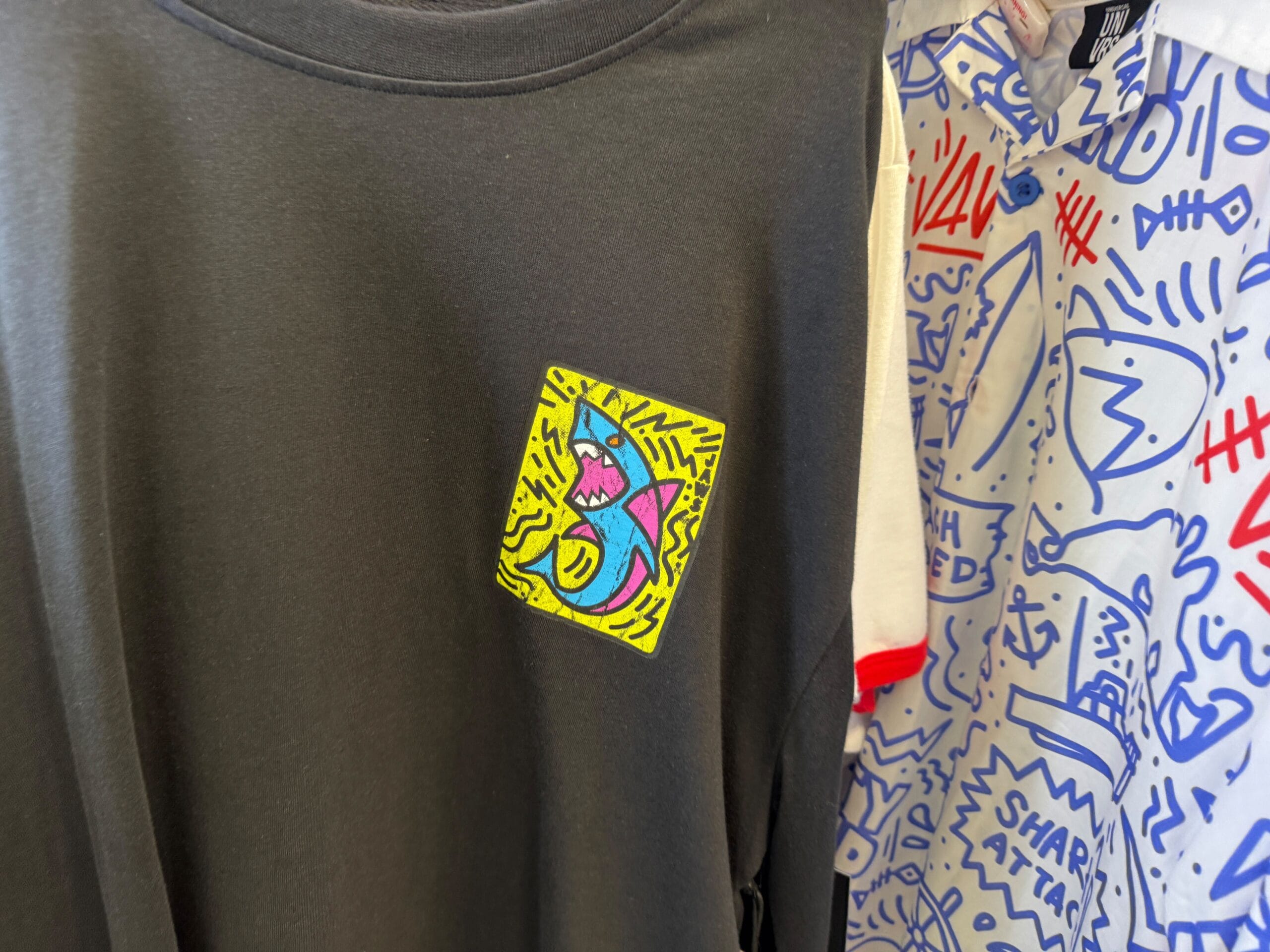 Black t-shirt with a colorful cartoon shark patch; whimsical doodle-patterned shirts beside it, like Disney gift shop displays.
