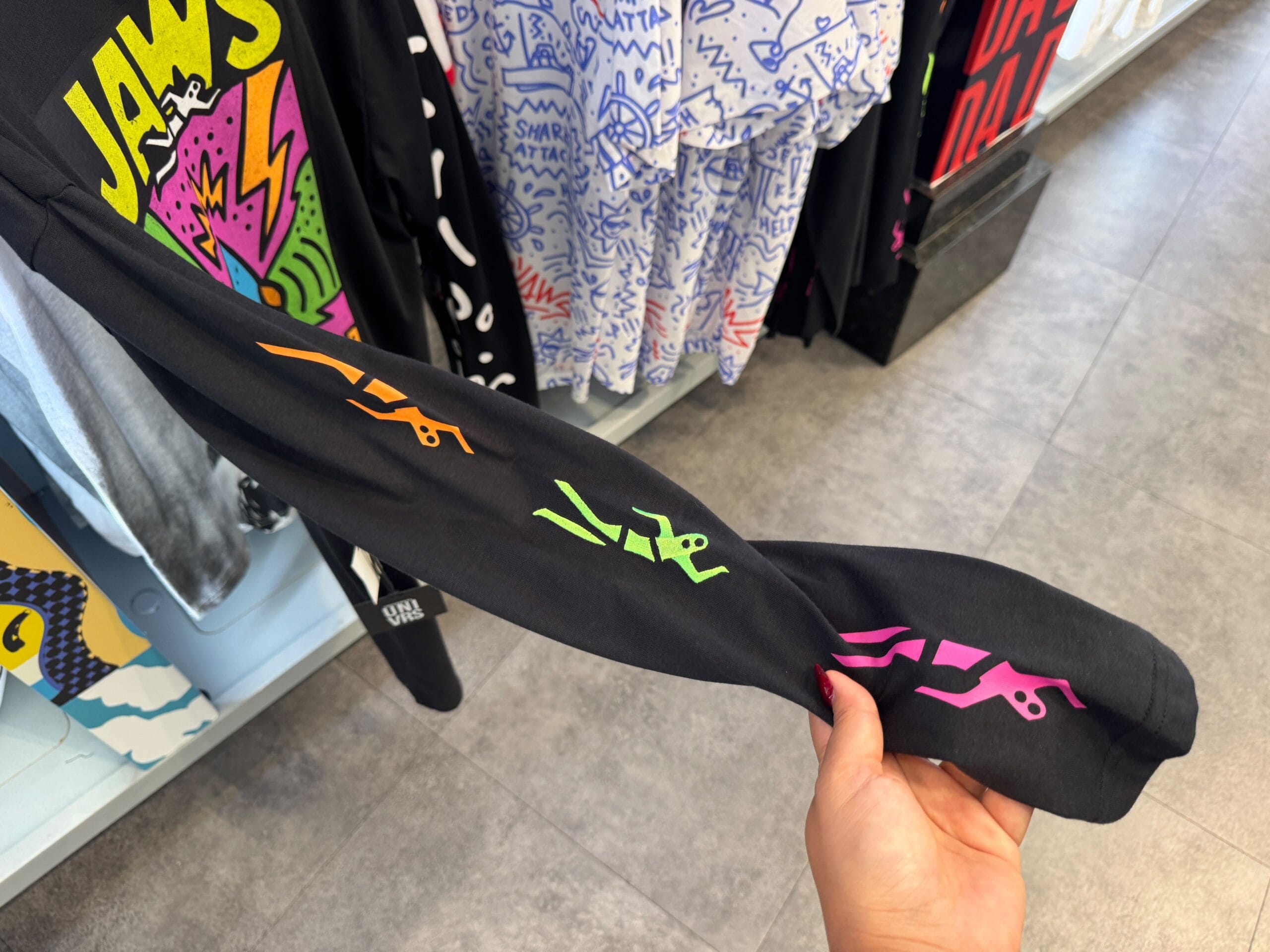A hand holds a black sleeve with colorful stick figures; behind, graphic shirts hang on a rack like those found at Disney parks.