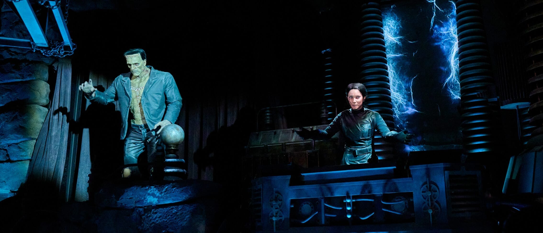 A Monsters Unchained Frankenstein stands by a lab as a woman works at a panel under blue light, no Disney castle in sight.