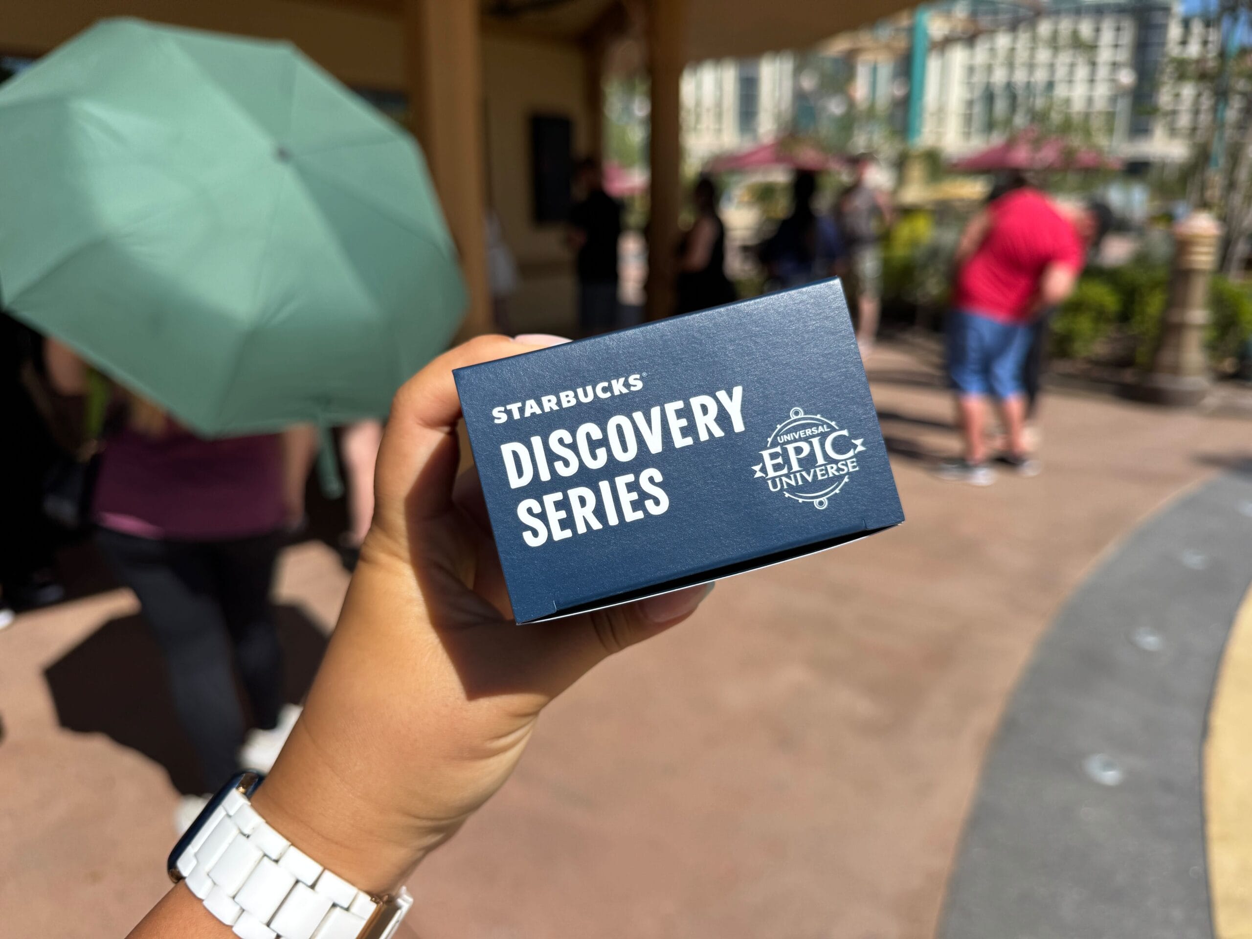 A hand displays a Starbucks Discovery Series Epic Universe box outdoors, with guests and an umbrella in the background—no castle visible.