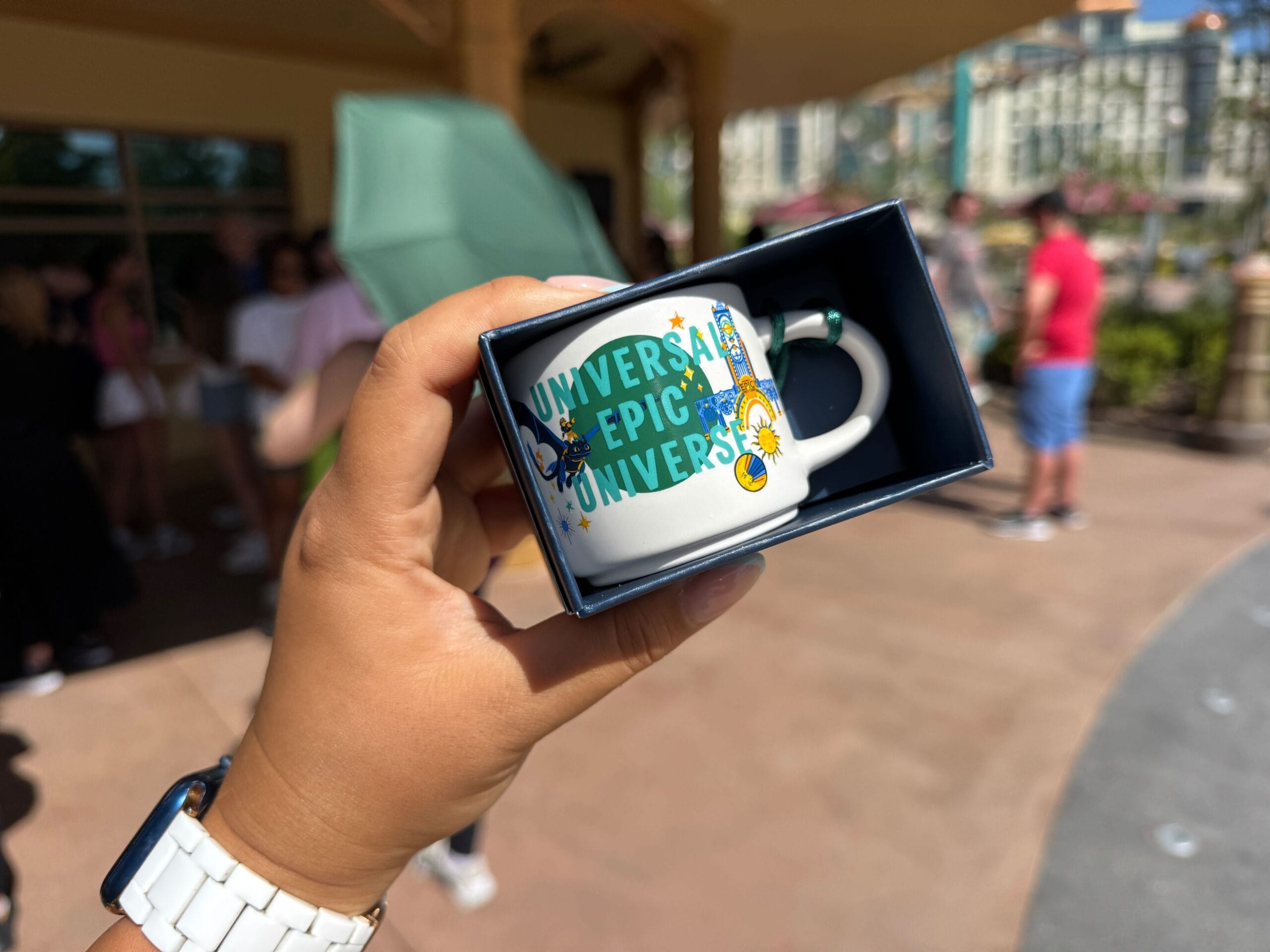 A hand holds a "Universal Epic Universe" mug in a box, with a theme park scene behind—no Disney castle like Cinderella’s present.