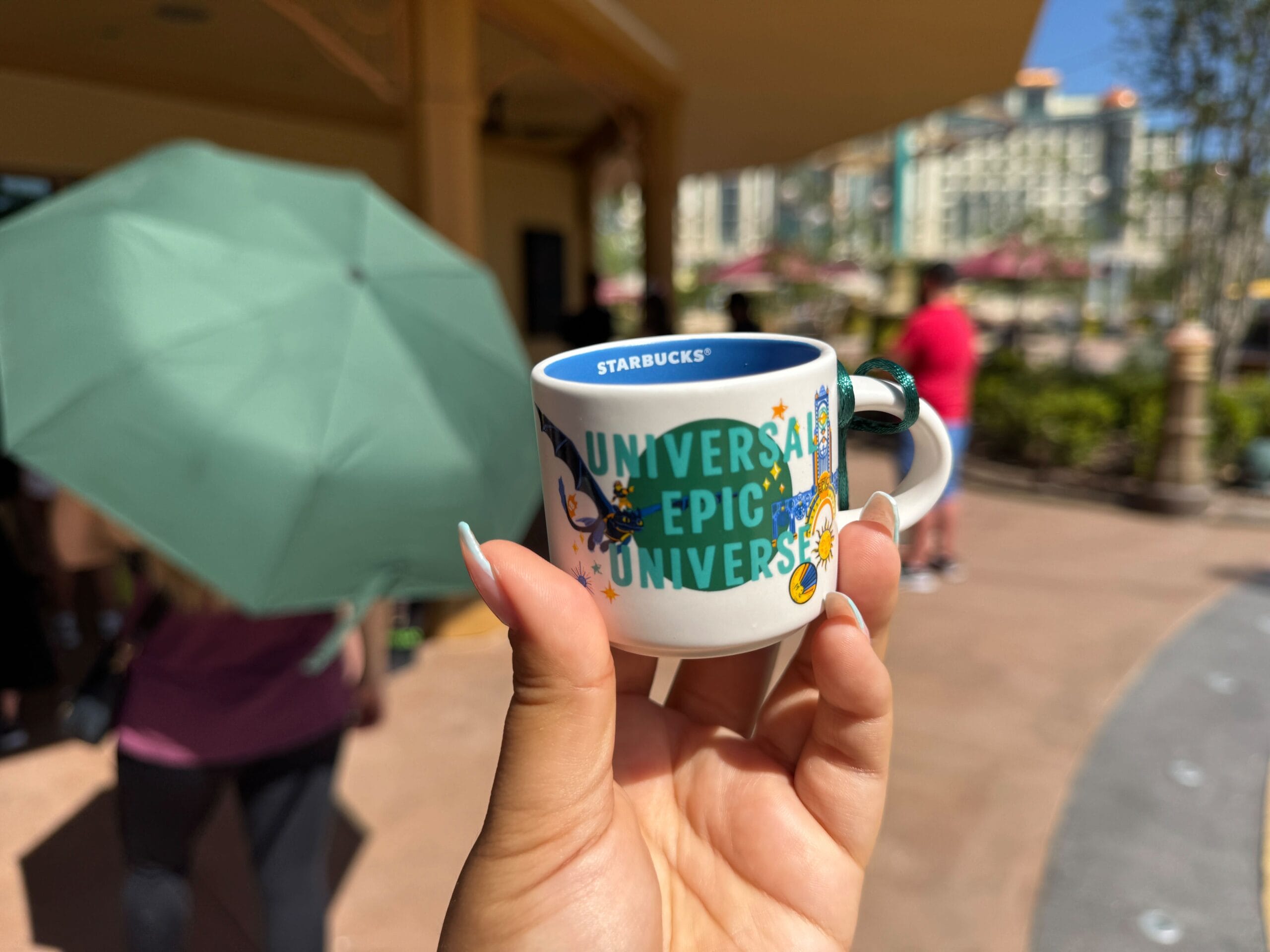 A hand holds a small Starbucks "Universal Epic Universe" cup outside on a sunny day, but no Disney castle is visible to compare.