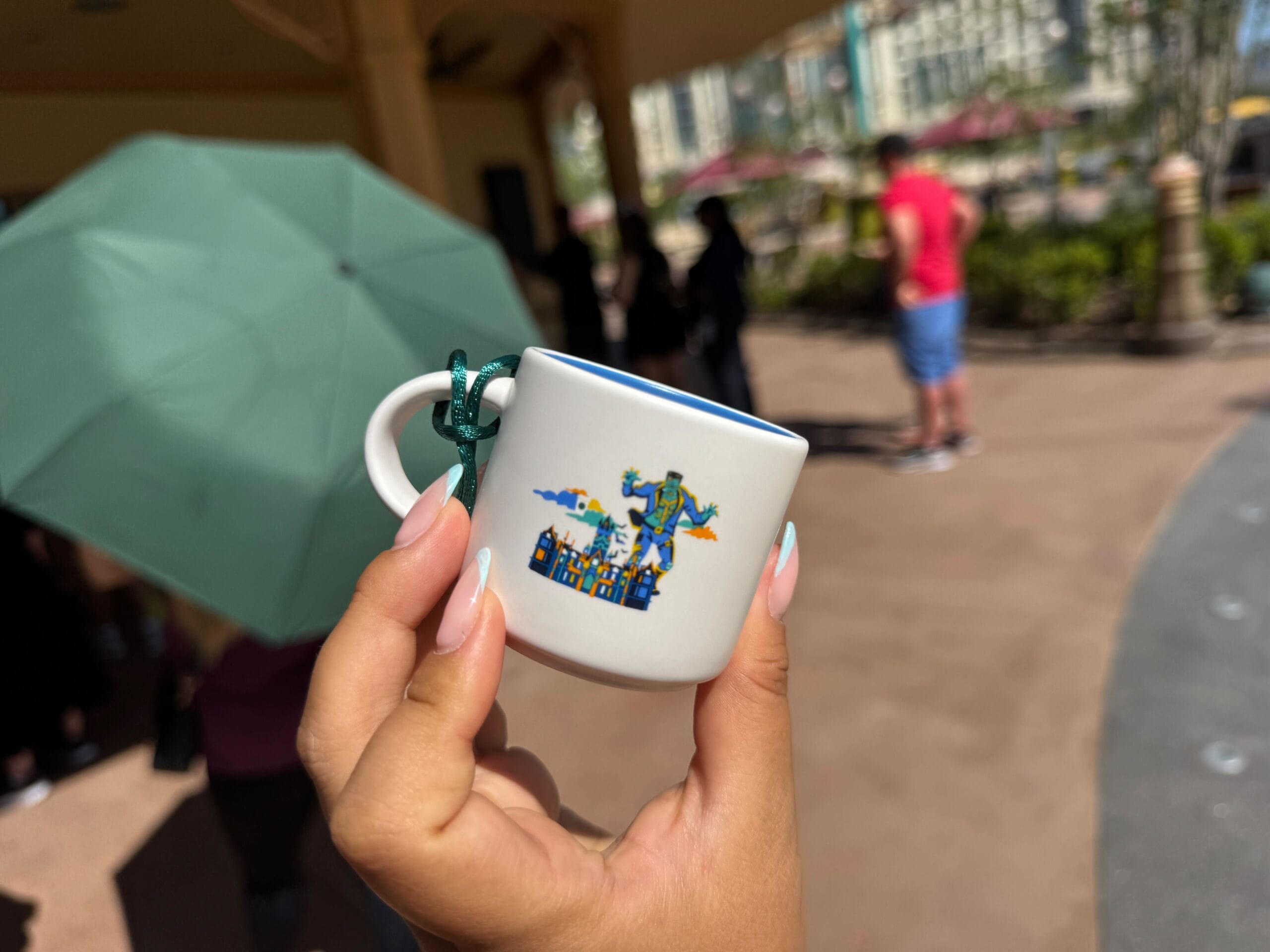 A hand holds a small souvenir cup with a vibrant city design, reminiscent of Disney parks merchandise; blurred guests and umbrella behind.