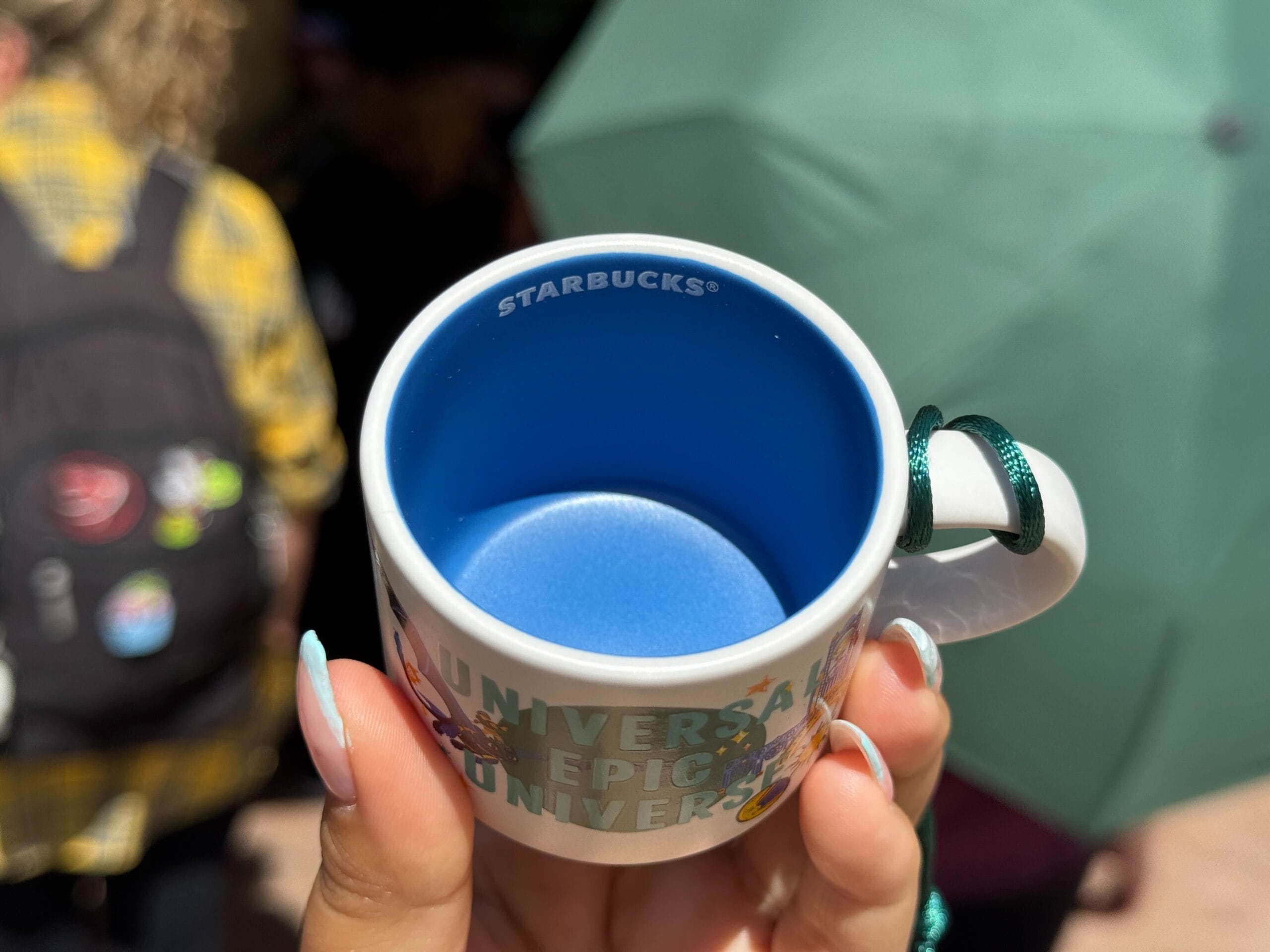 A hand holds a small Starbucks cup with a blue interior; in the background, park guests and a signature Disney green umbrella are visible.