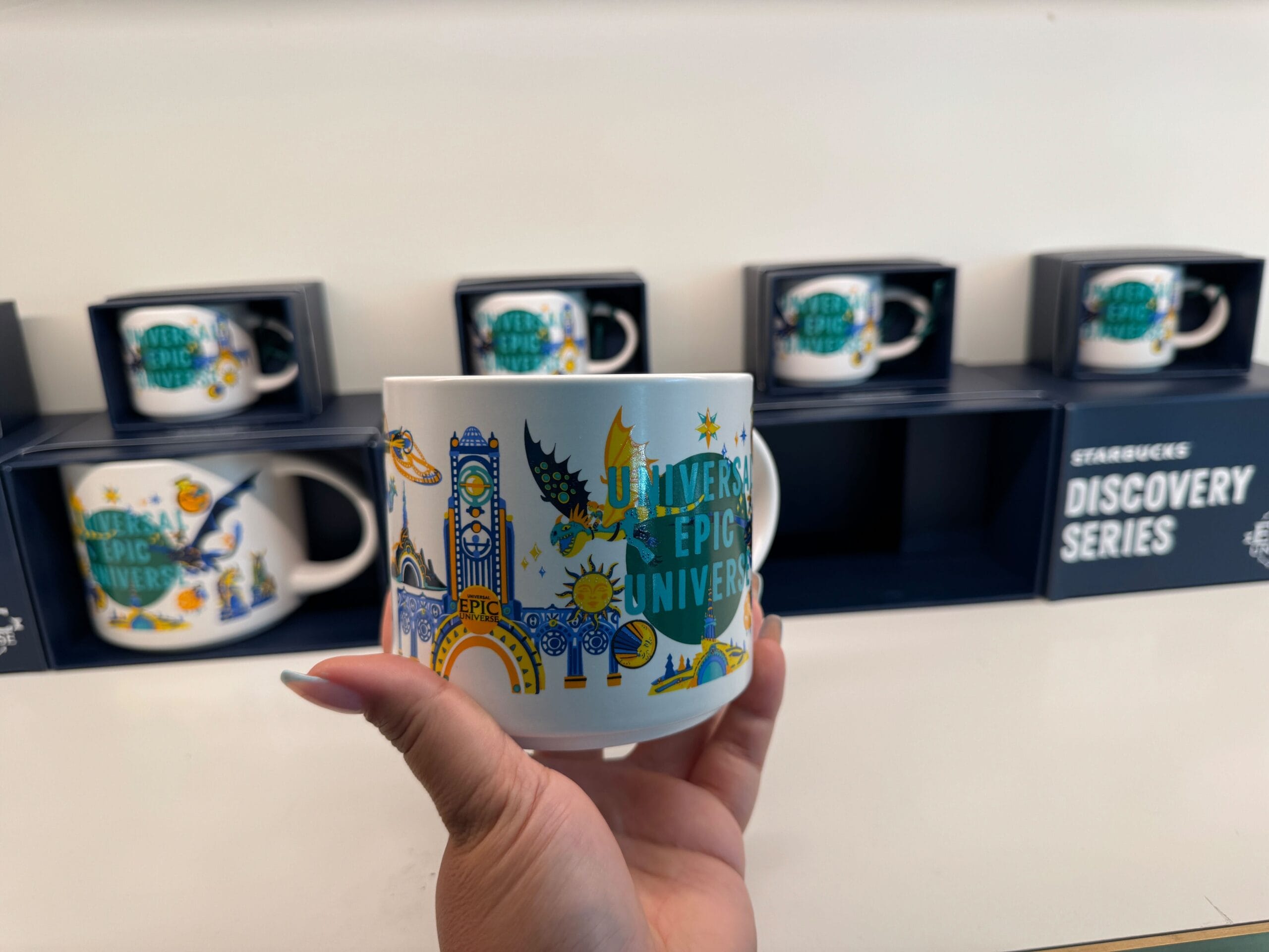 Hand holding a Starbucks mug with a vibrant Universal Studios design; other mugs in boxes behind, not featuring Disney park castles.