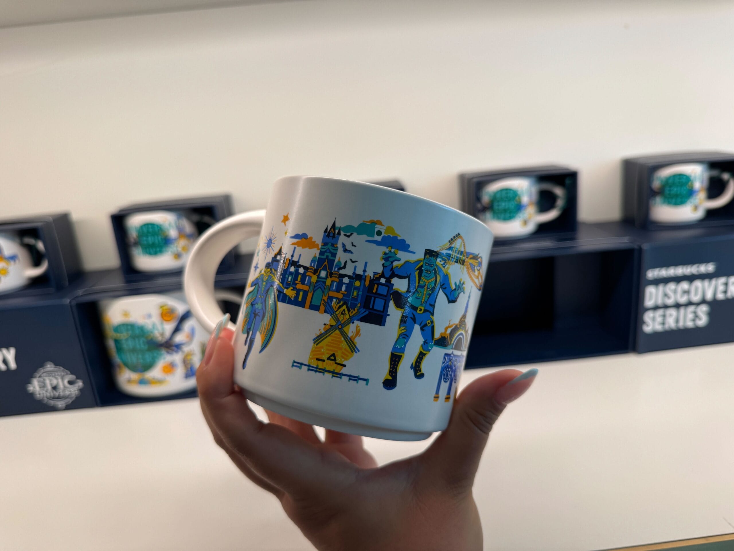 A hand holds a Starbucks mug with vibrant city and knight art, resembling Disney parks designs; more mugs, like park collectibles, behind.
