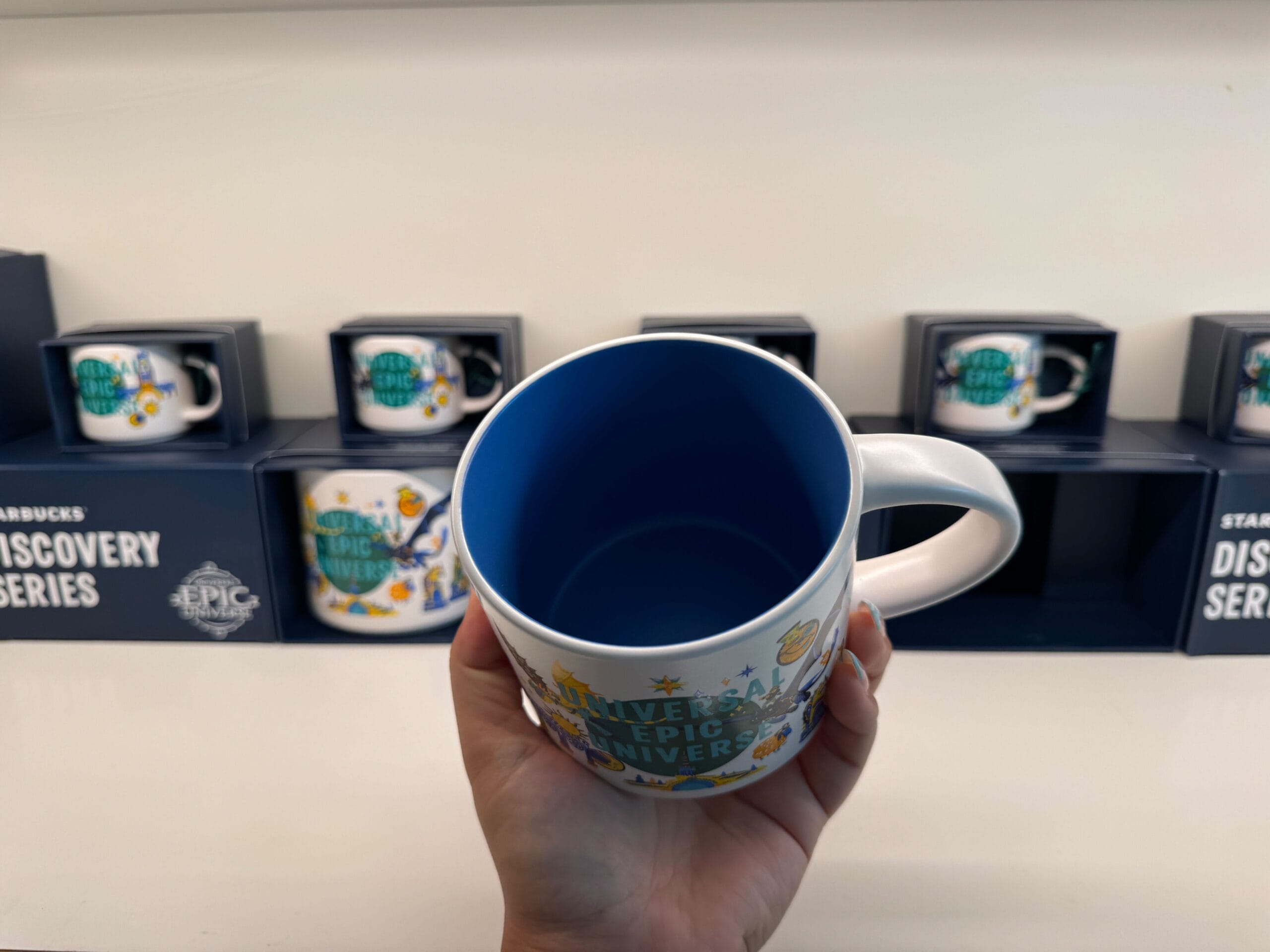A hand holds a blue-and-white Starbucks mug in front of boxed mugs on a display shelf, similar to those found in Disney parks.