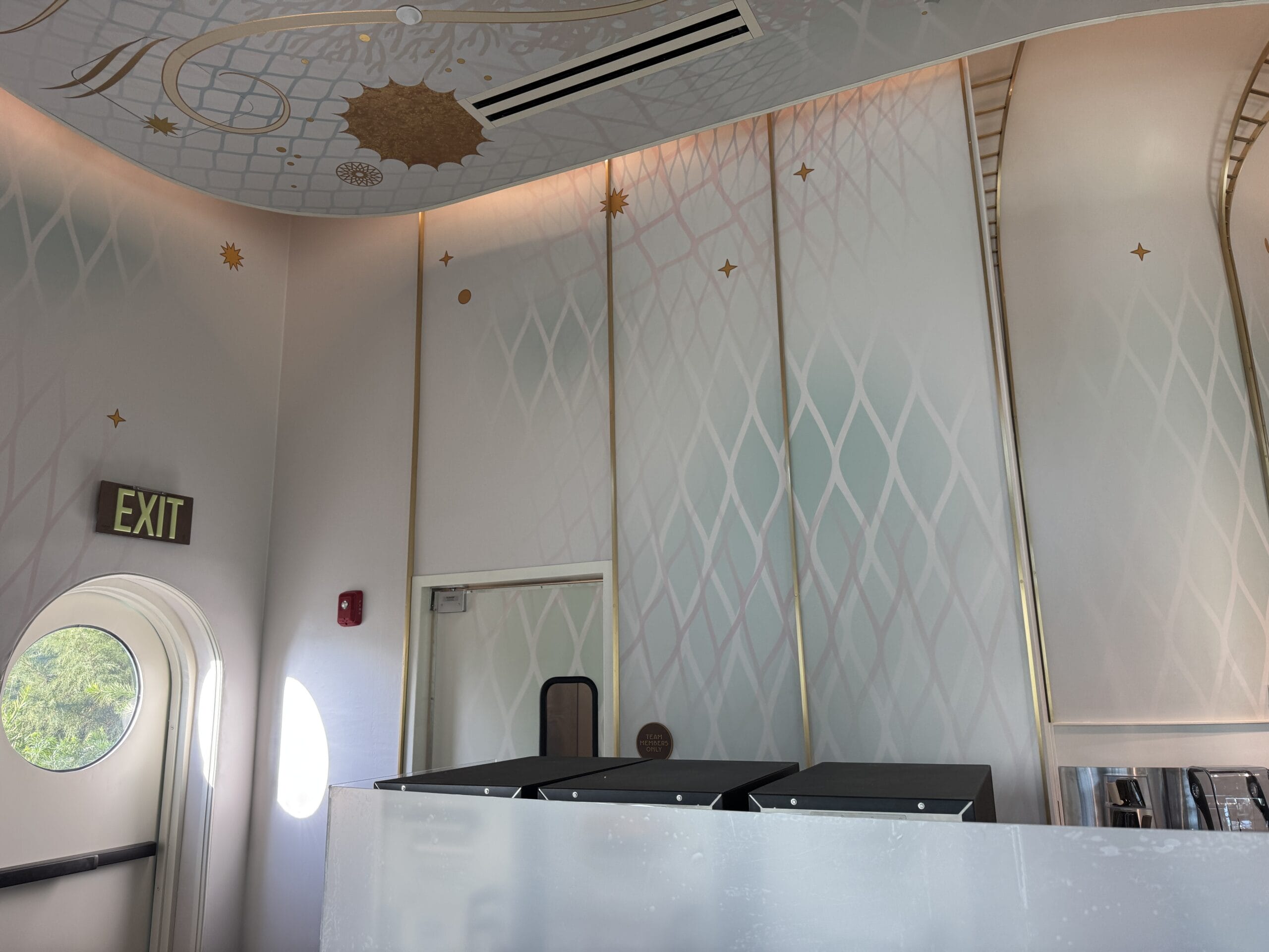 A pastel room with gold accents, Starbucks trash bins, an exit sign, and a left-side door, much like Disneyland’s Main Street style.