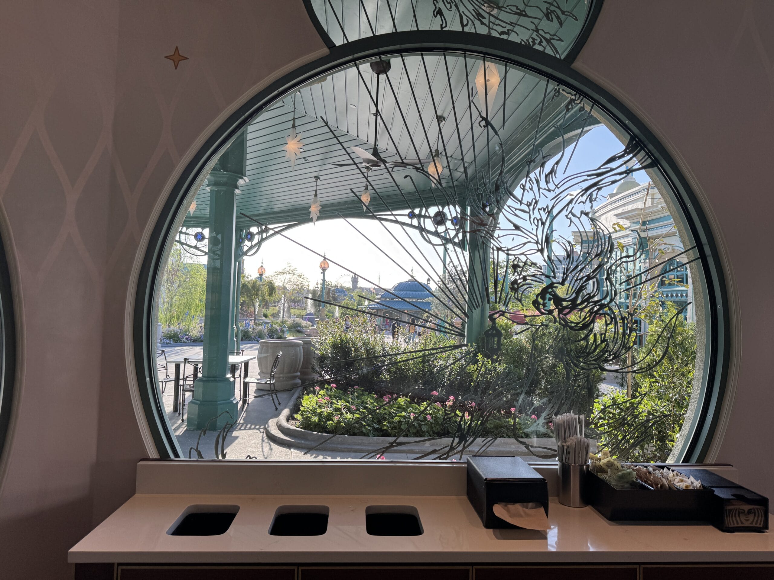 A large round window with etched designs overlooks a garden and teal patio outside Starbucks, reminiscent of Disney Park elegance.