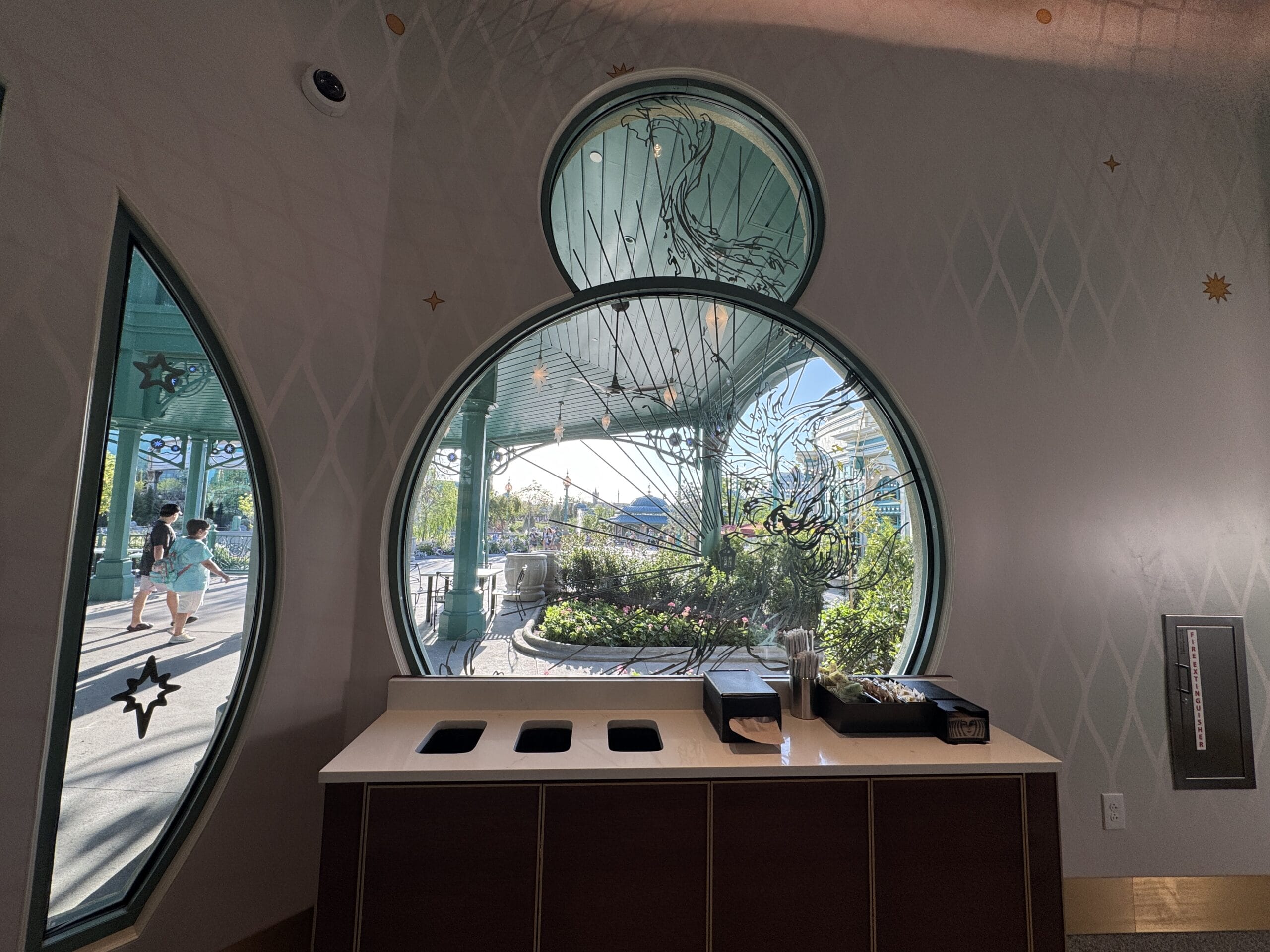 Sunlight streams through grand round windows behind a Starbucks waste station at Epic Universe, reminiscent of Disney park architecture.