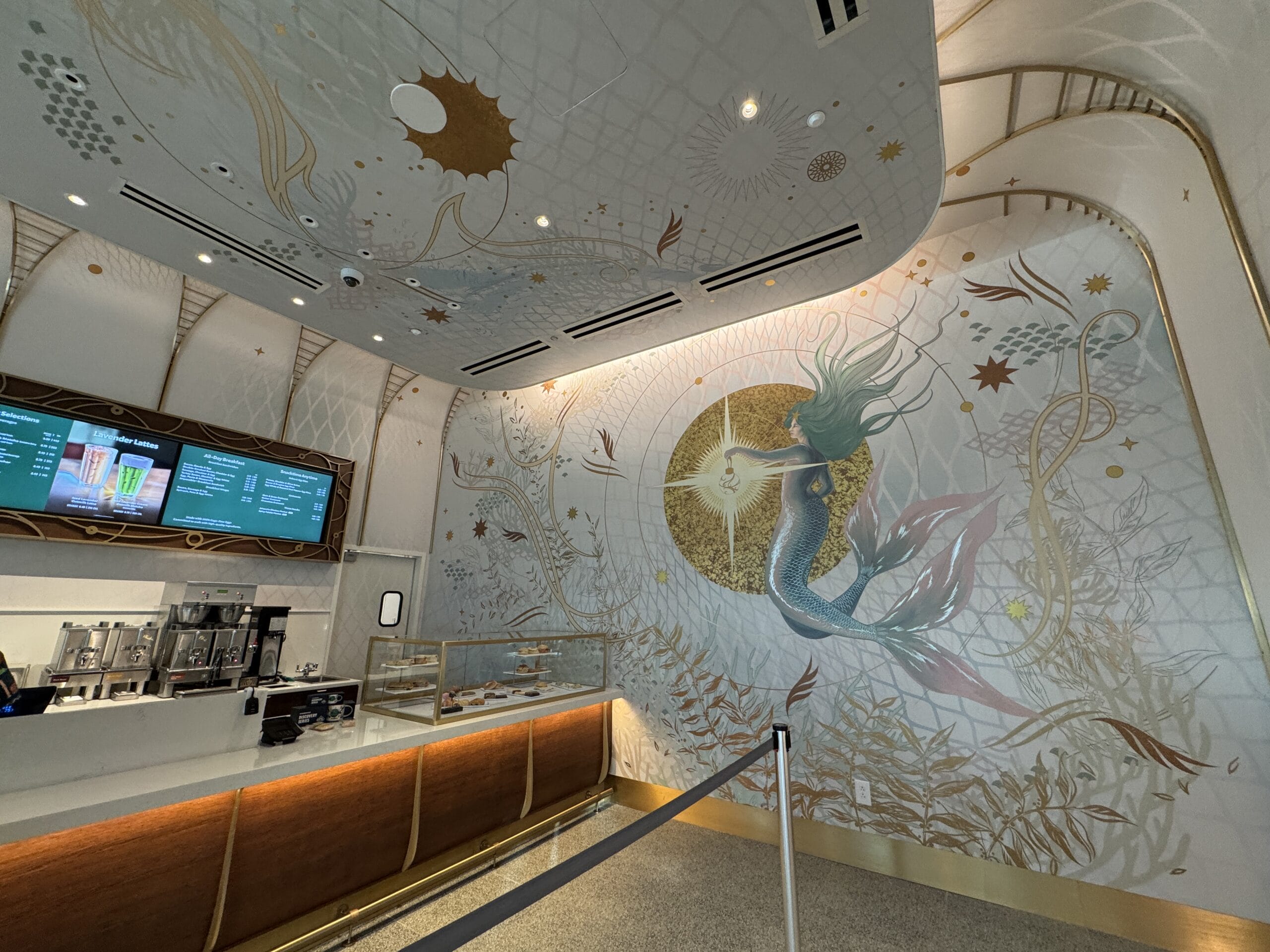 Inside a Starbucks cafe, a large mermaid mural and sea-themed gold decor evoke a whimsical vibe, reminiscent of Disney park artistry.