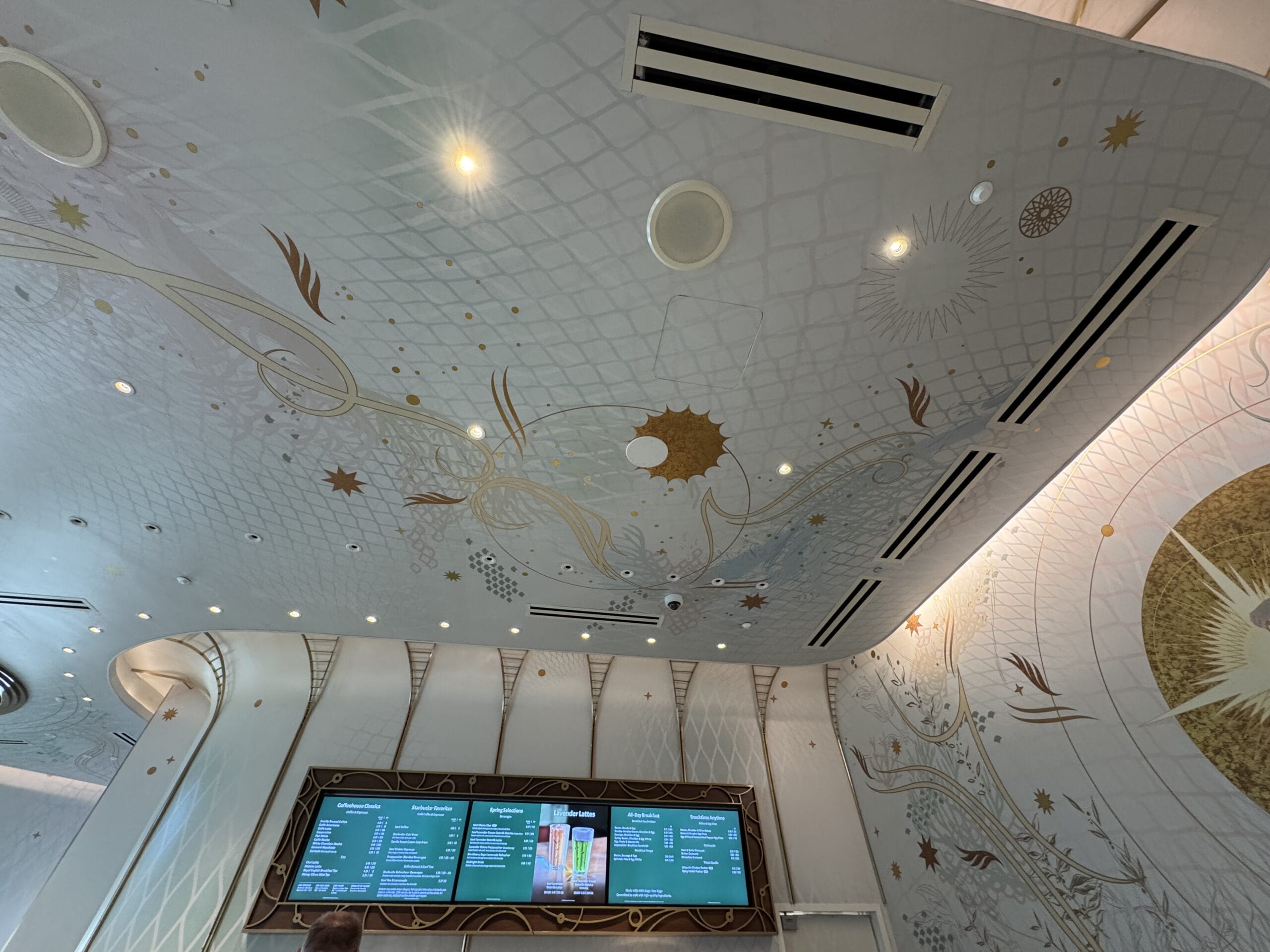 Elegant white and gold ceiling with celestial motifs, reminiscent of Disney park details, above a menu in a modern café setting.