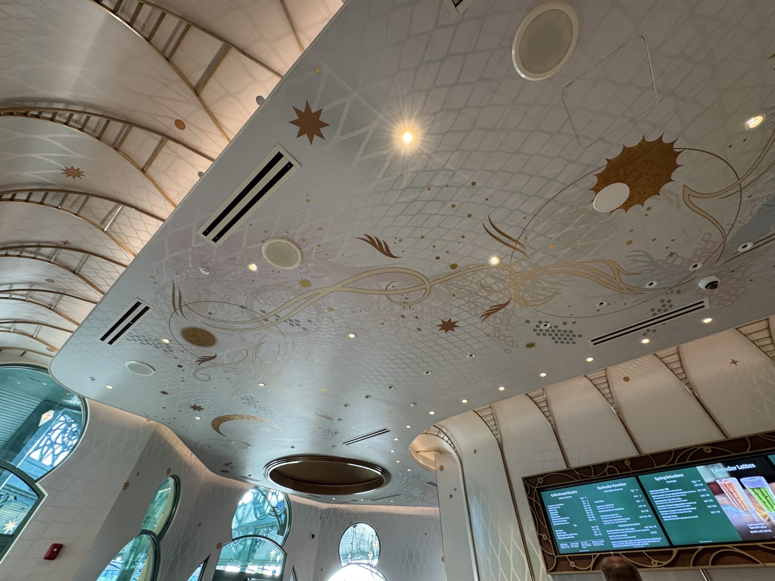 Elegant white ceiling with gold stars and lights tops a Starbucks at Epic Universe, echoing Disney castle grandeur above arches.