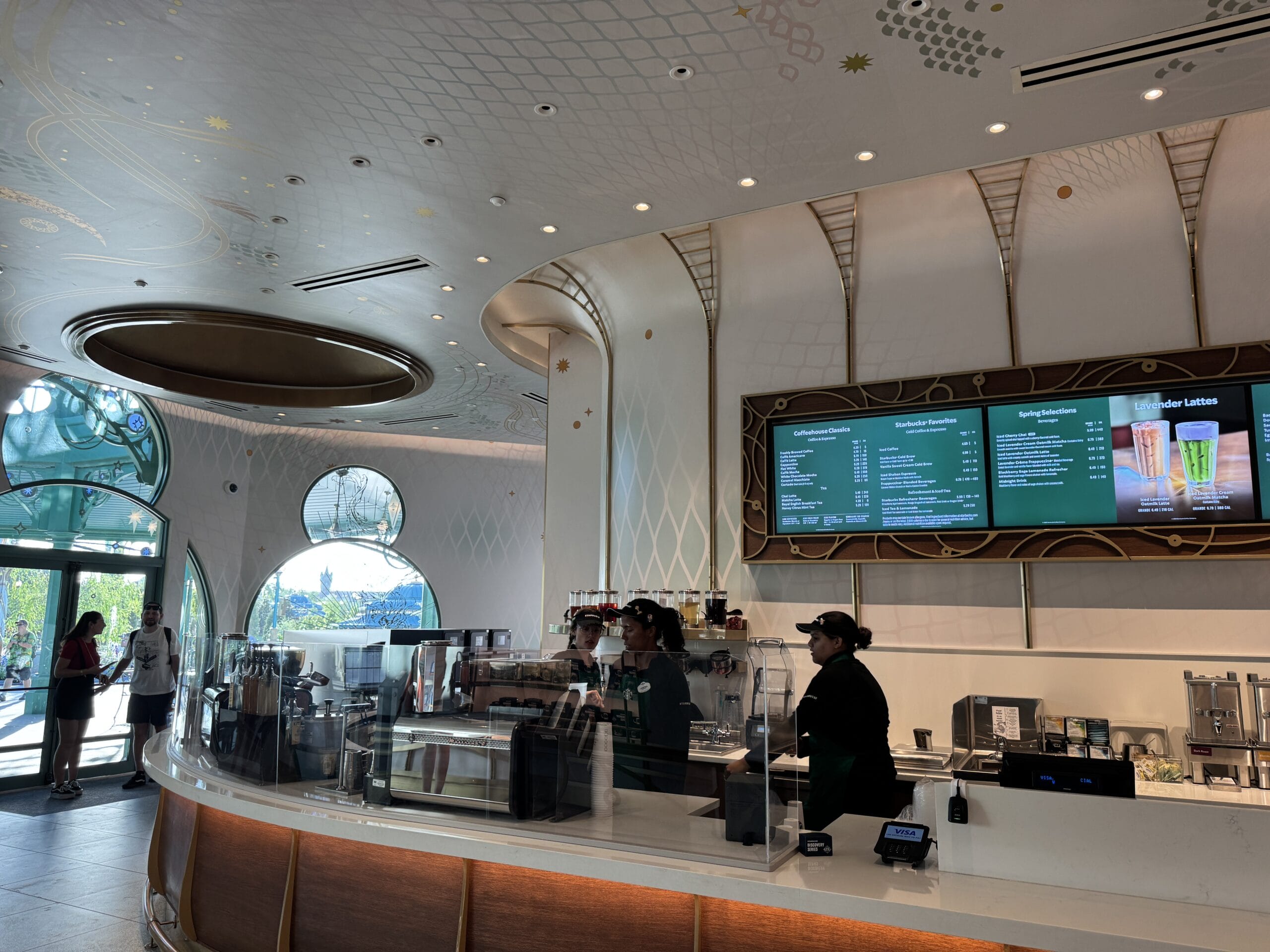 Trendy coffee shop at a Disney park, staff behind counter, digital menus above, guests near big windows with park views outside.