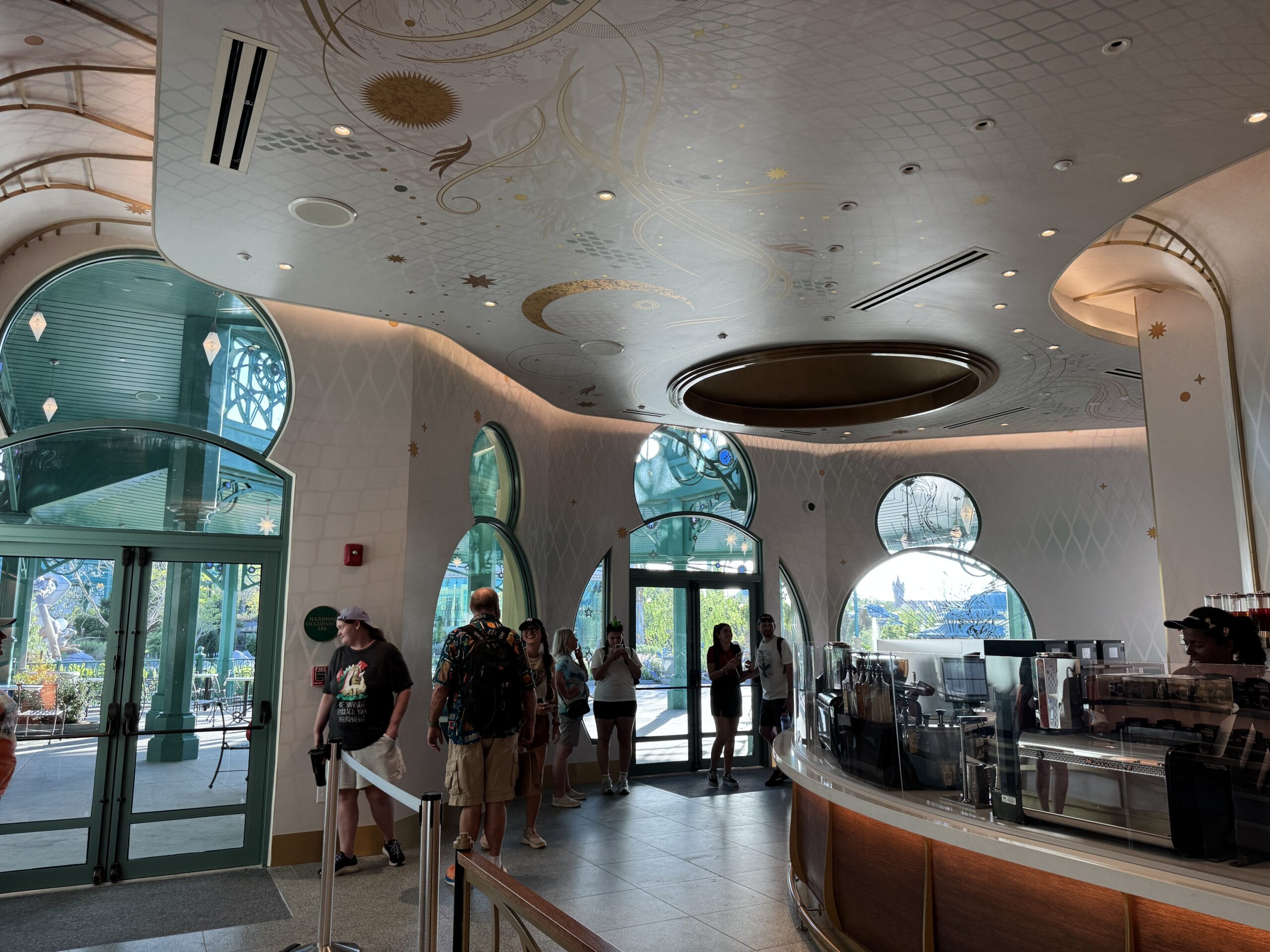 Guests gather inside a modern Starbucks at a Disney park, sunlight streaming through large windows beside the curved counter.