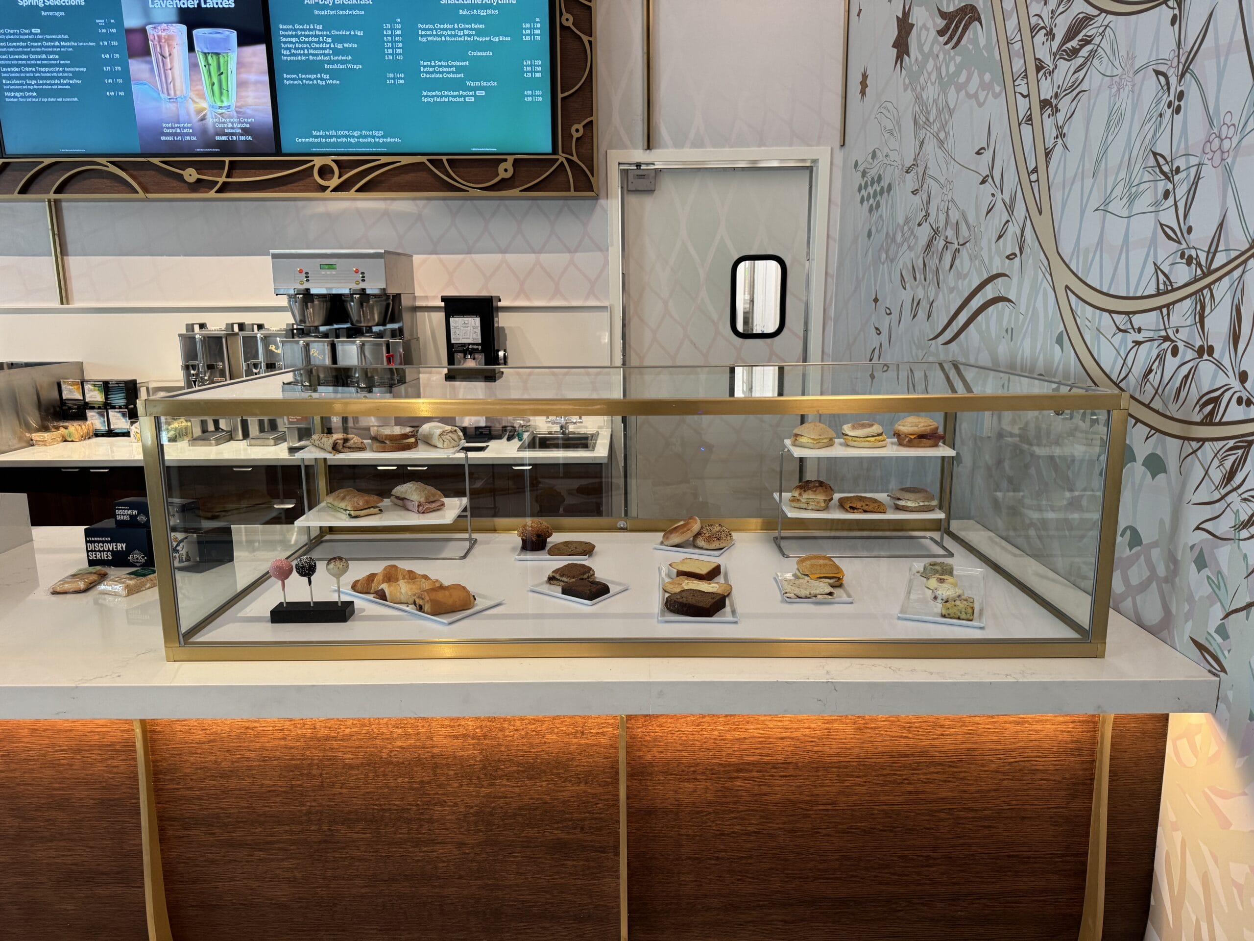 Glass bakery case with assorted pastries and sandwiches in a stylish café, similar to Starbucks at Disney Parks, no castle visible.